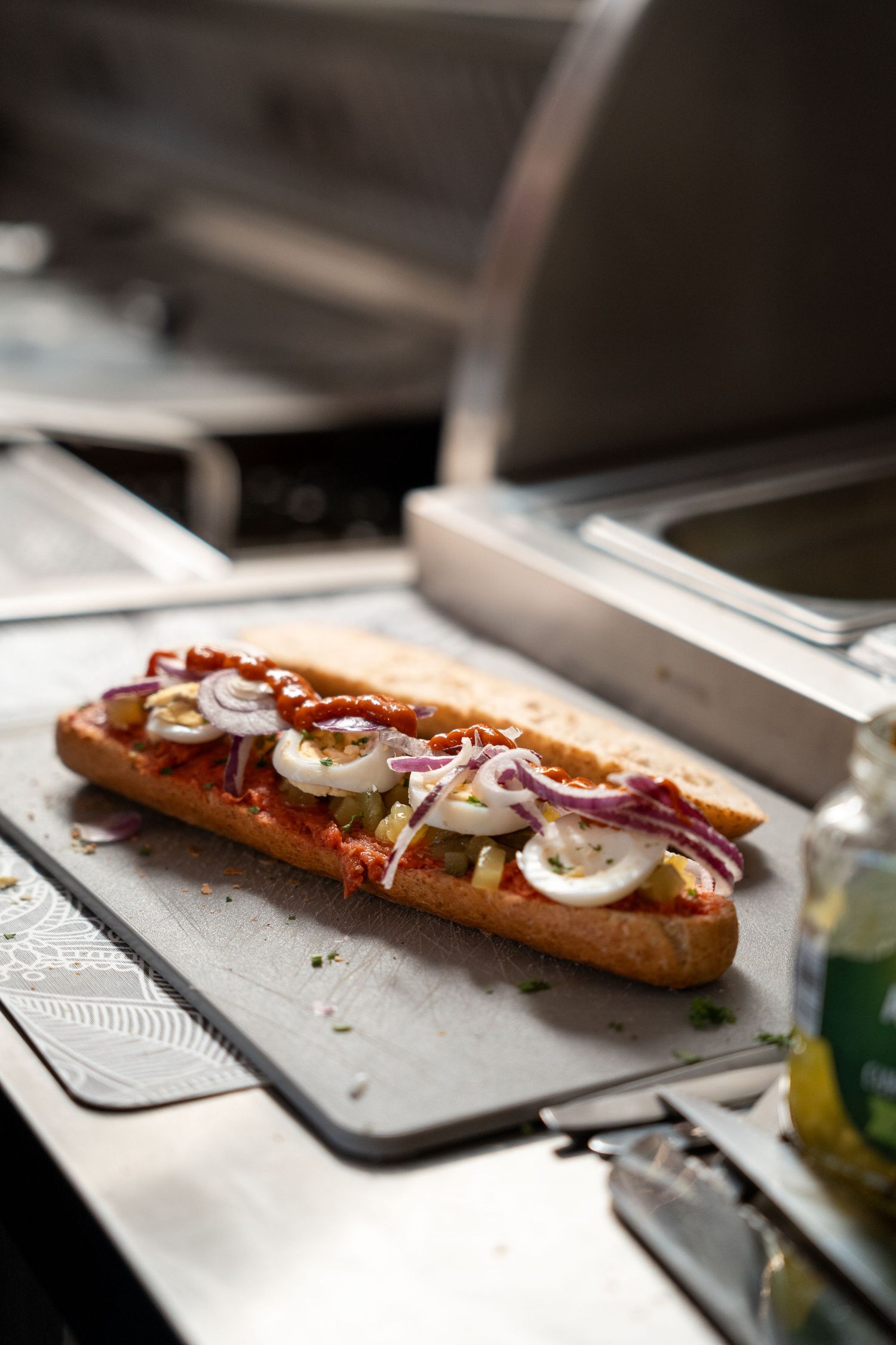 Open-faced baguette sandwich topped with tomato sauce, sliced eggs, red onions, and olives on a metal tray.