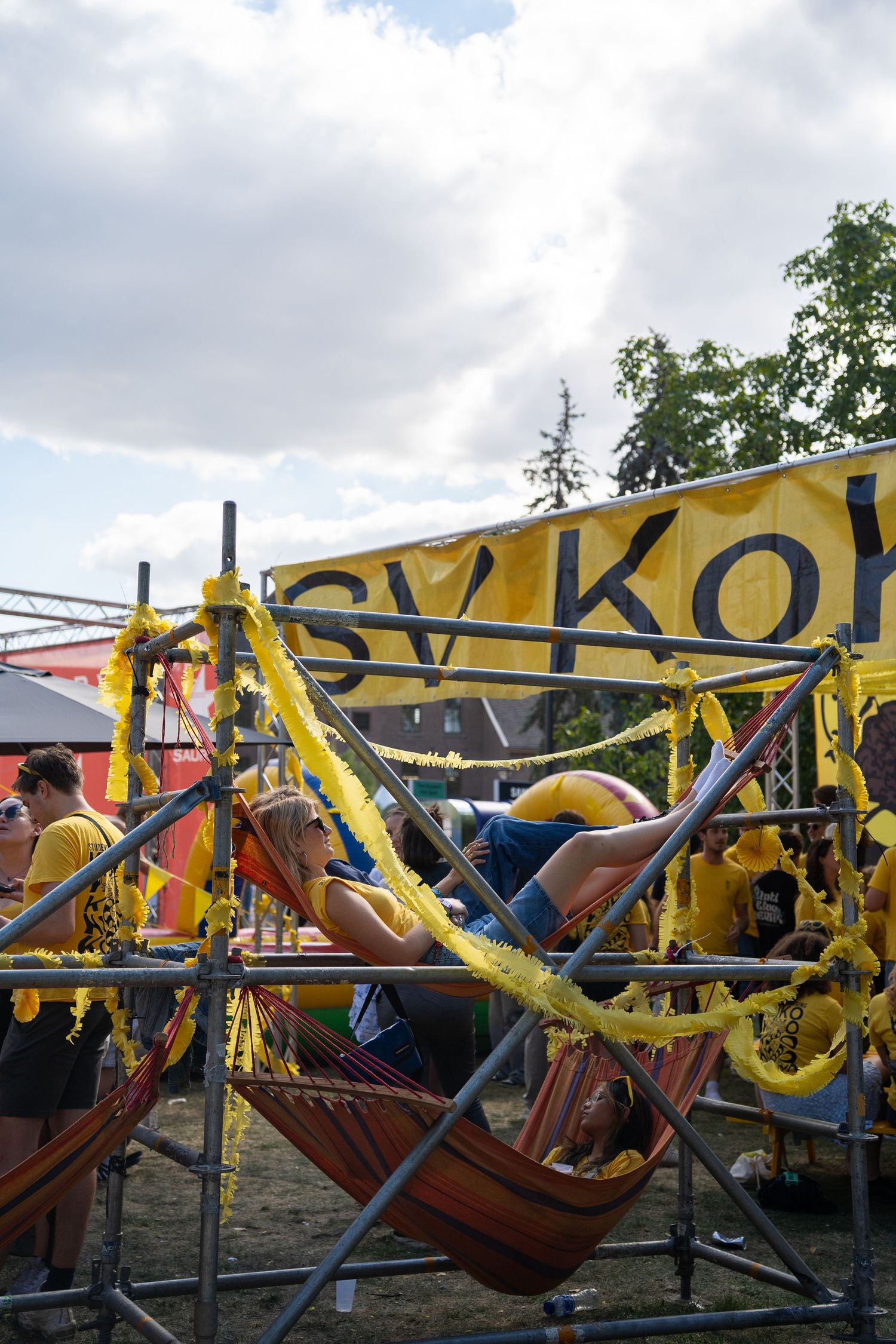 Person relaxing in a colorful hammock at an outdoor festival with yellow decorations and a banner in the background.
