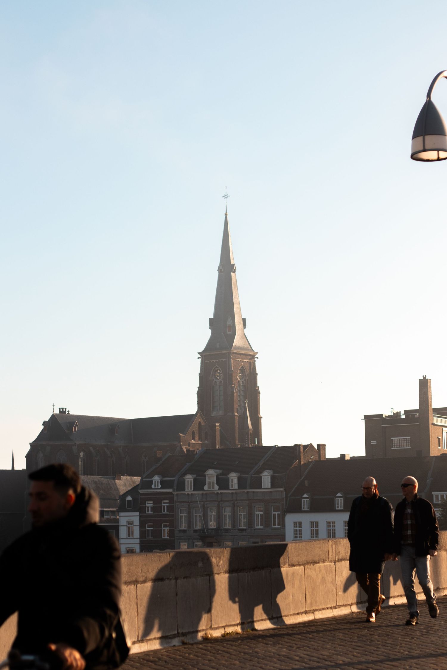 Pedestrians casting long shadows on a bridge with a tall Gothic church spire rising above rooftops.