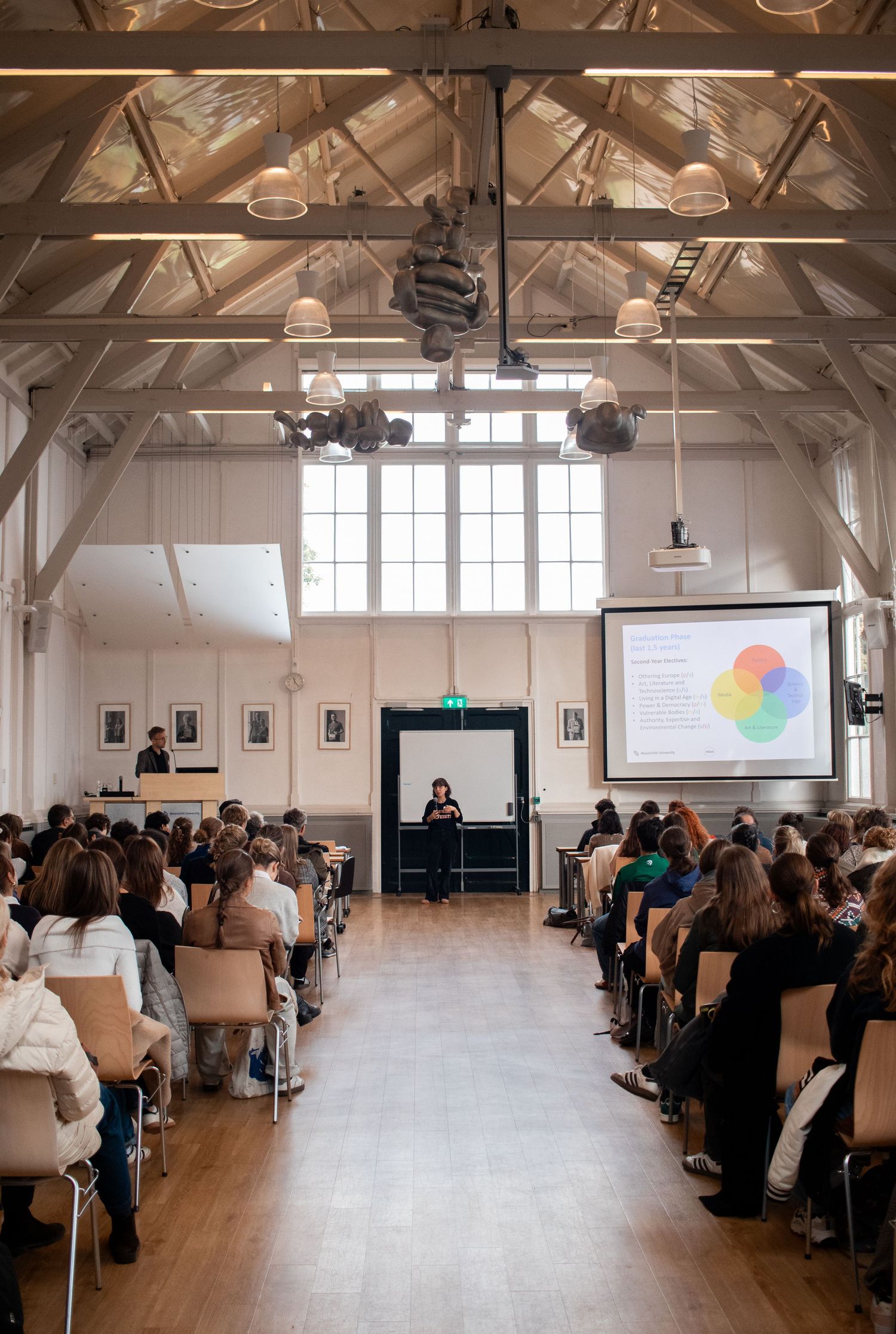 Large audience attending a presentation in a bright white hall with vaulted ceiling and projected slides.