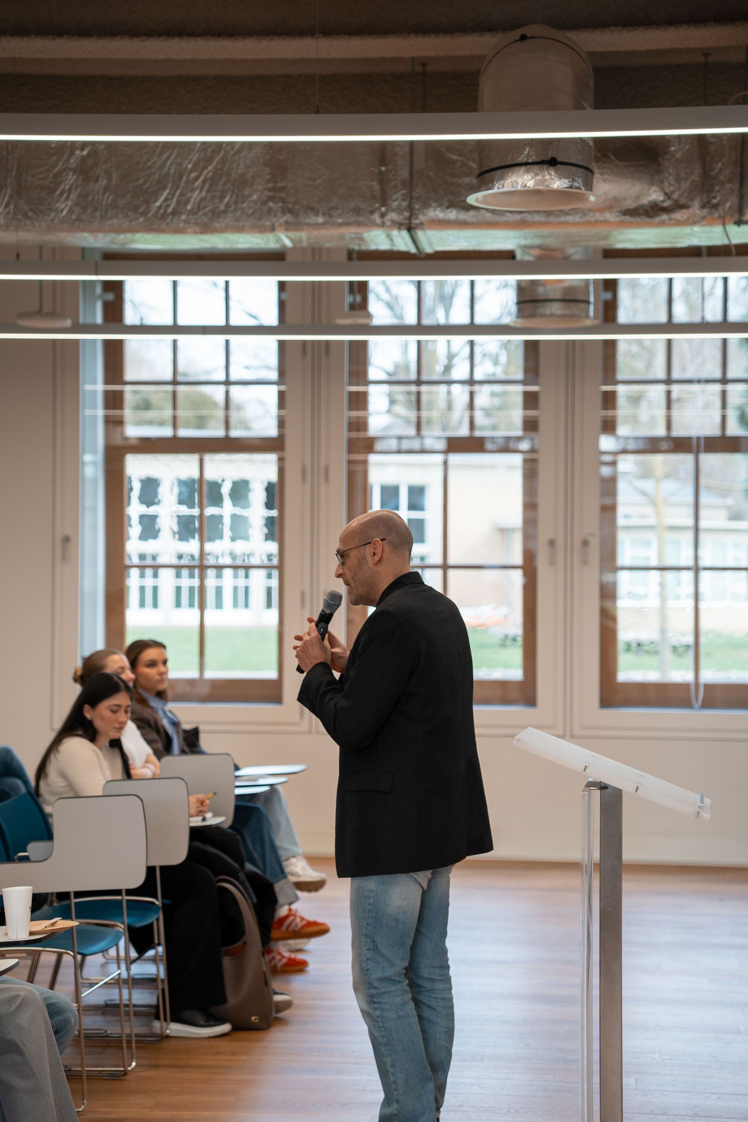 Bald man in black blazer speaks into microphone at indoor academic presentation with seated audience.