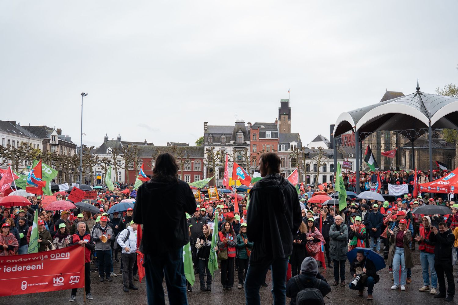 Large crowd of protesters with colorful flags and banners gathered in a town square on a rainy day.