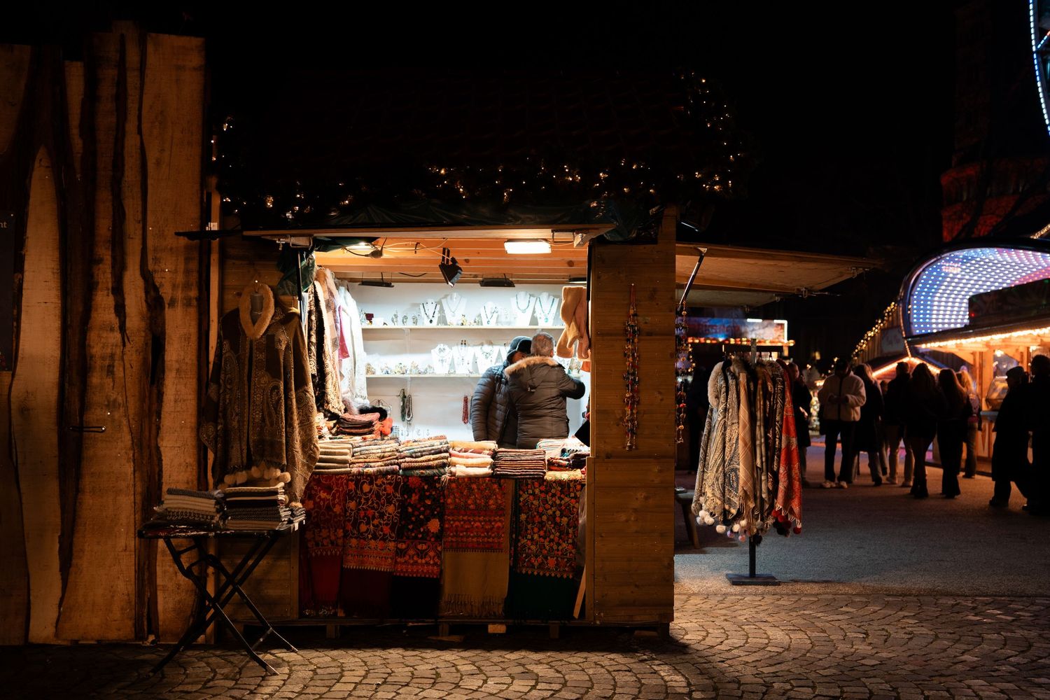 Cozy outdoor Christmas market stalls selling textiles and crafts on cobblestone streets at night.