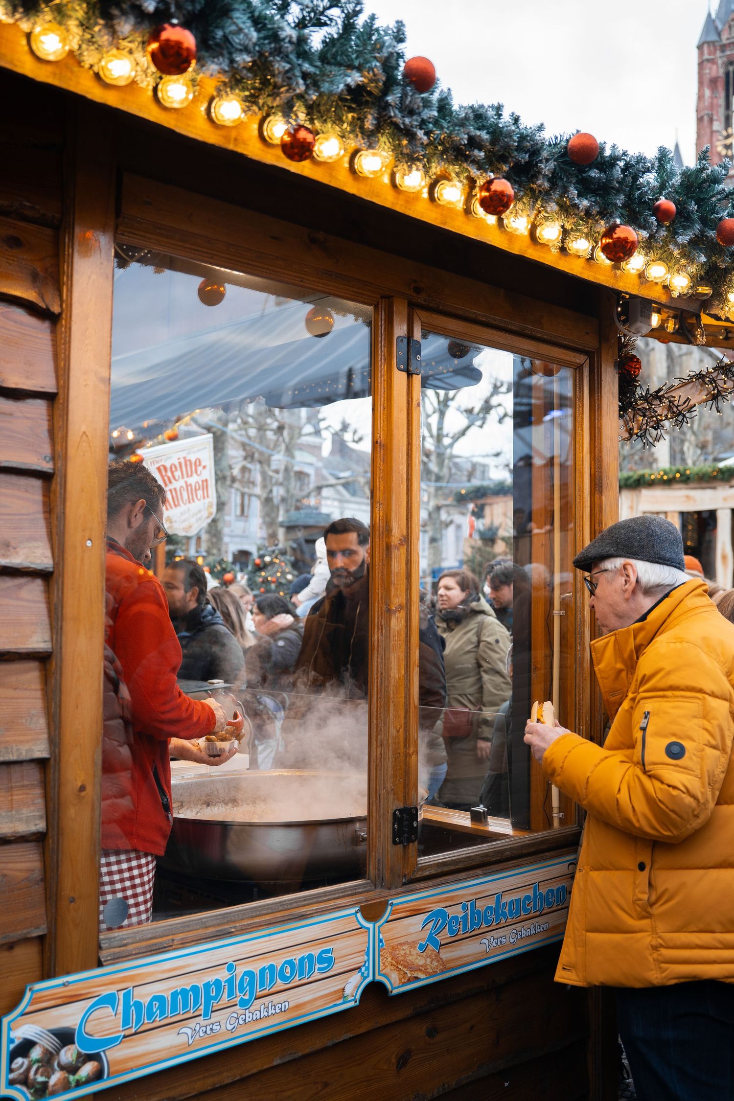 Man in yellow jacket eating at a steaming Christmas market stall selling champignons and Reibekuchen.