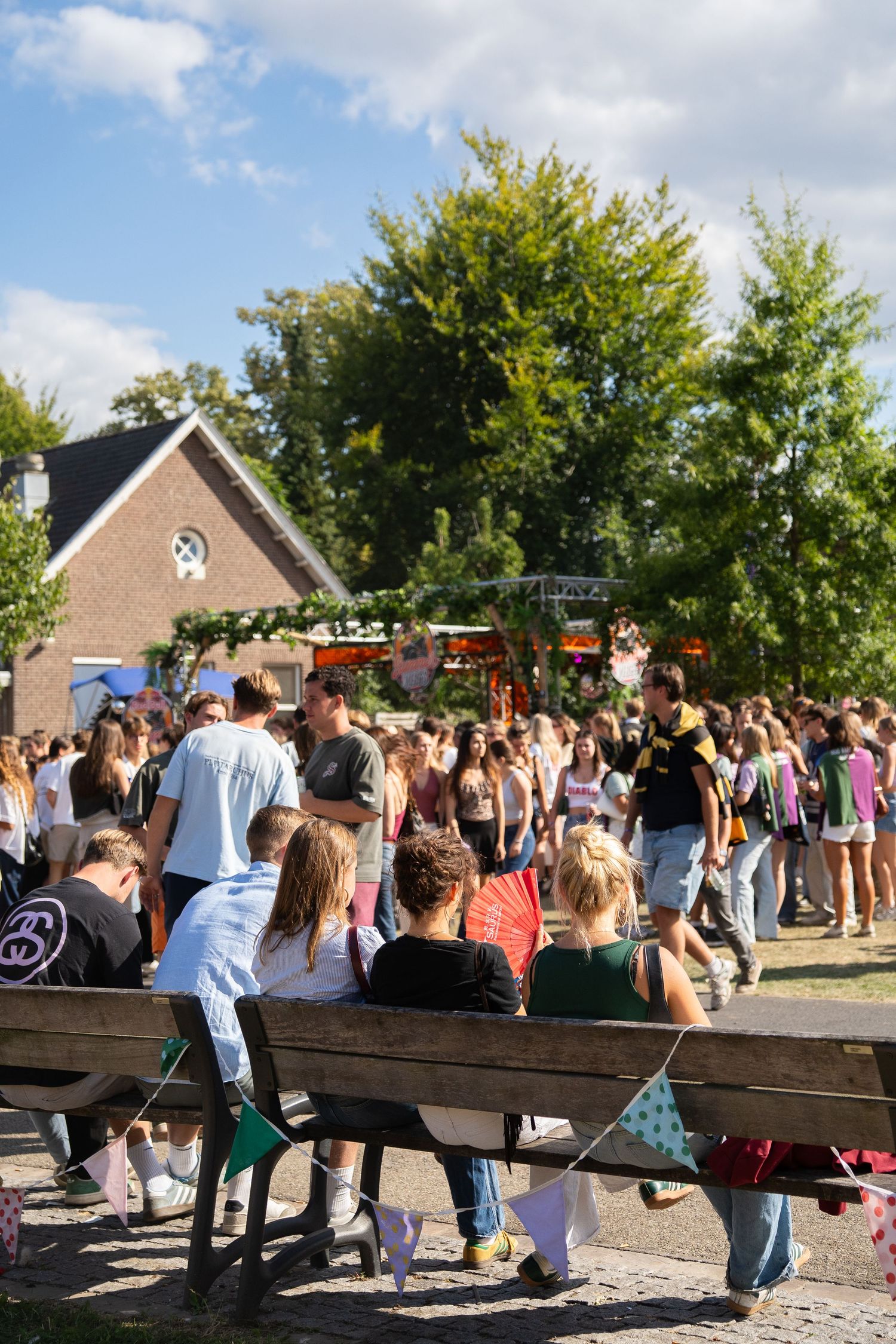 Large crowd of people gathered at a lively outdoor summer festival near a brick building and trees.