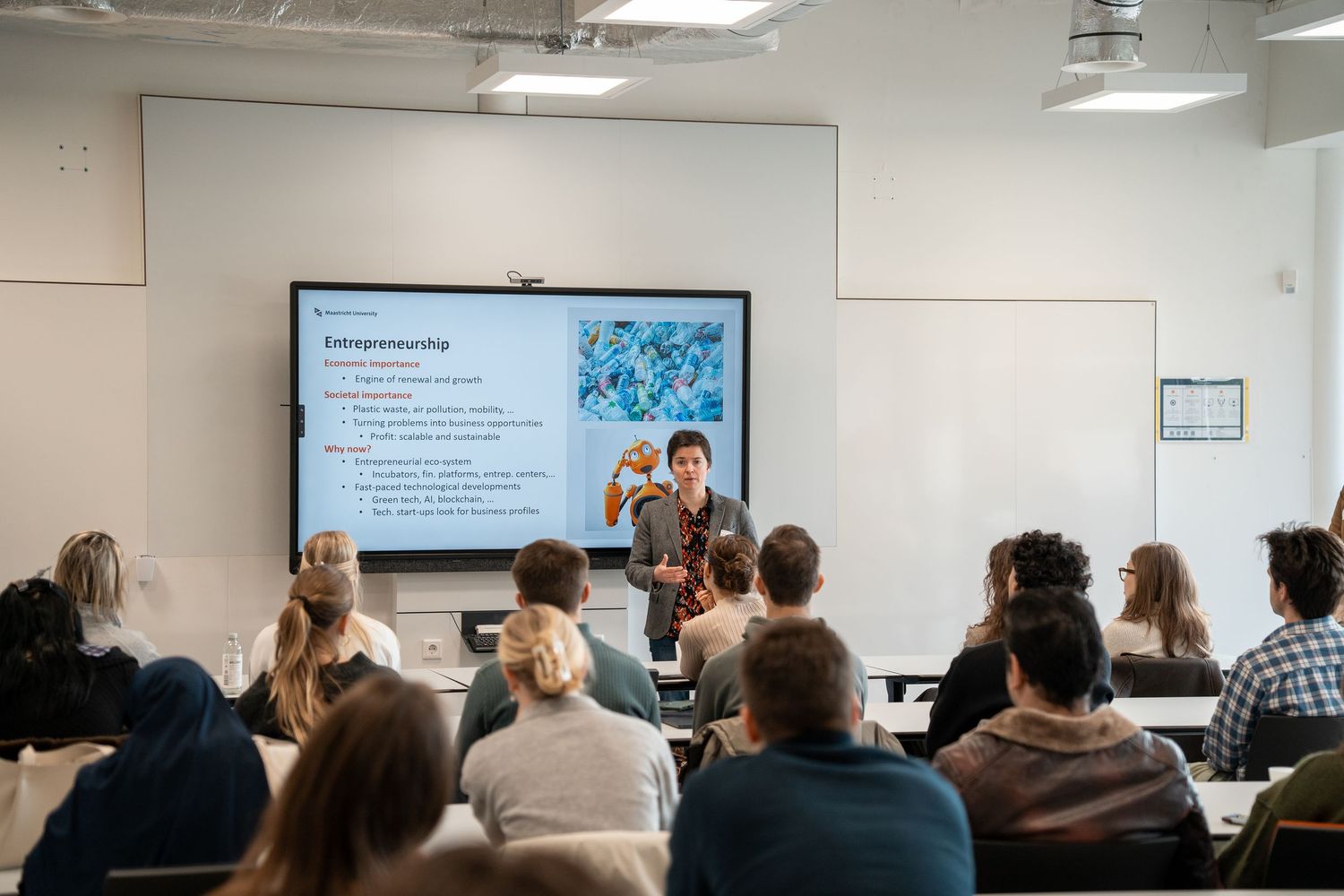Students attend an entrepreneurship lecture in a modern classroom with a presentation projected on a large screen.