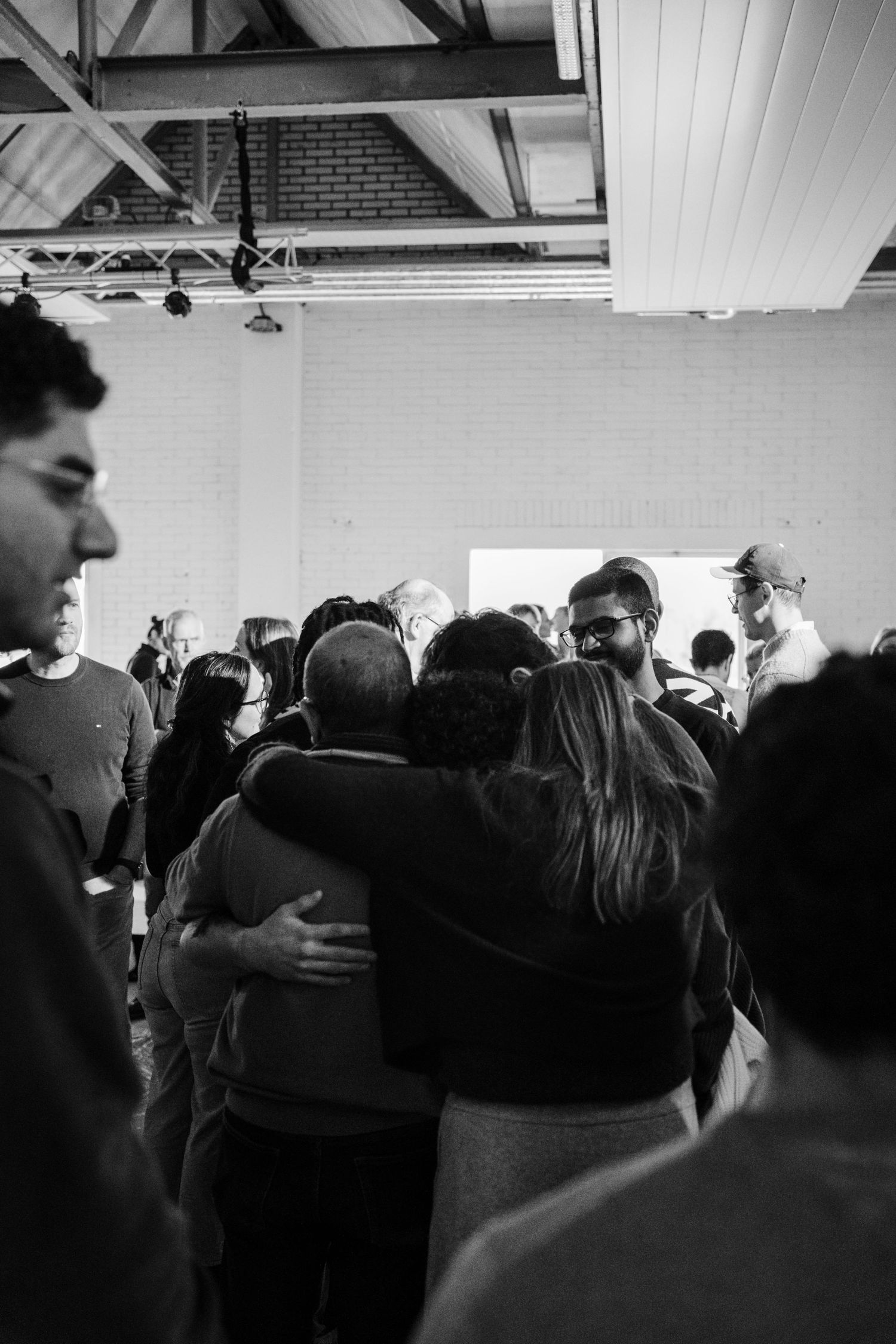 Black and white photo of people embracing in a warm group hug at an indoor community gathering.