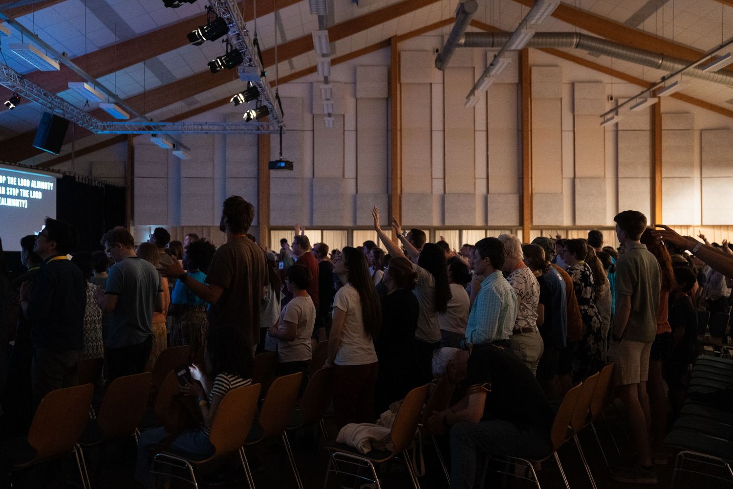 Congregation raising hands in worship inside a wooden-beamed church hall during evening service.