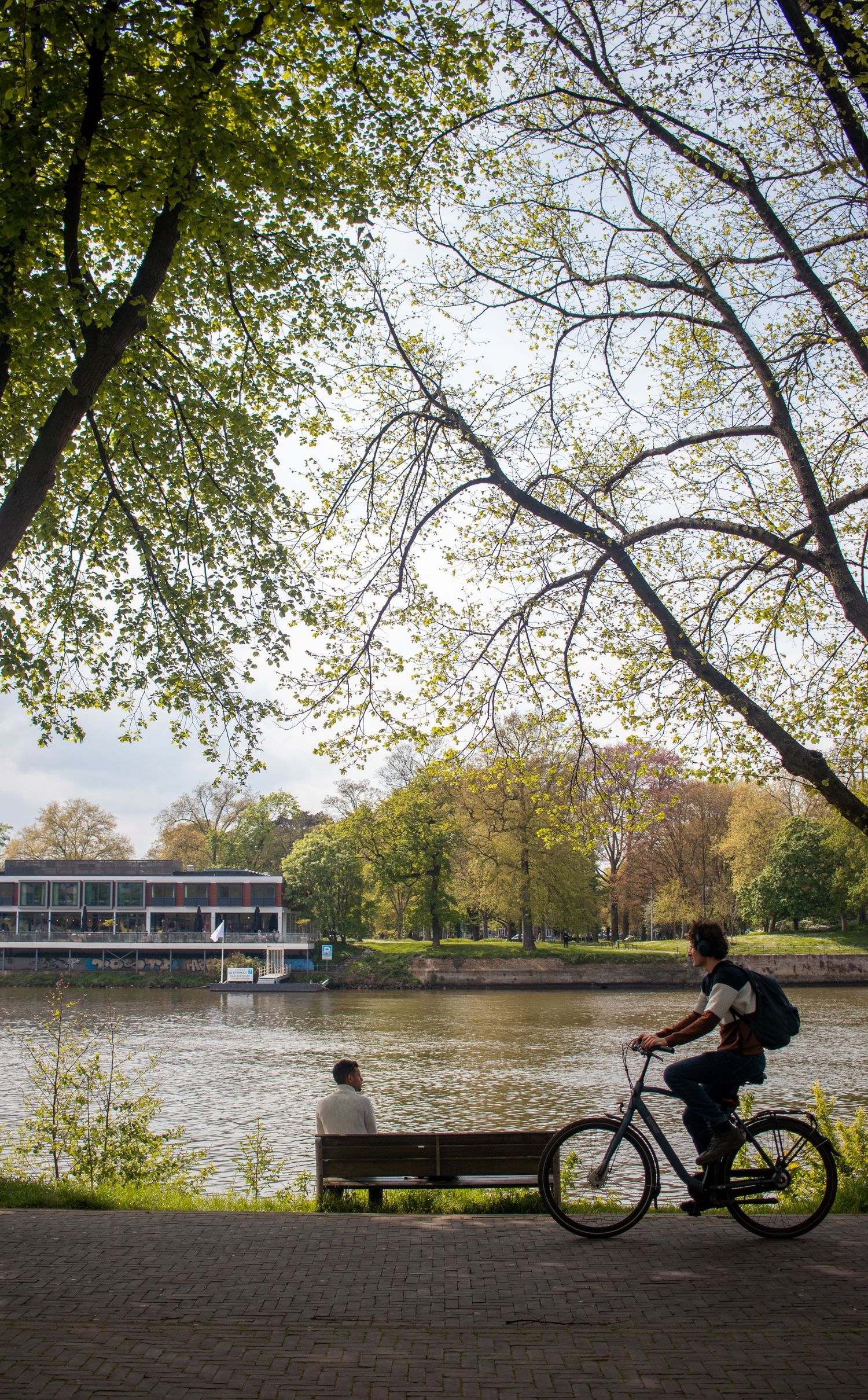 Cyclist riding along a scenic riverside path in a green park with a person sitting on a bench.