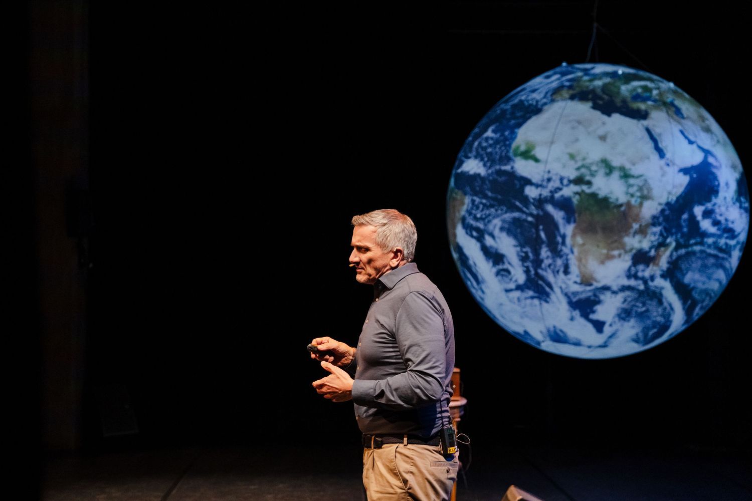 Male speaker presents on stage beside a large illuminated globe Earth projection in theater.