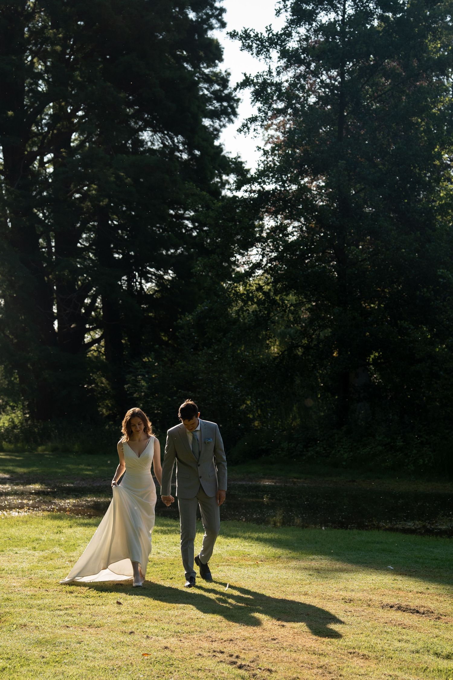 Bride in white gown and groom in grey suit walking hand in hand across sunlit green lawn near trees.