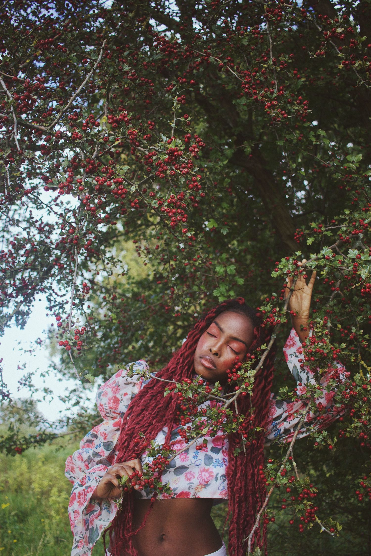 Woman with long red braids stands beneath a hawthorn tree with red berries in a lush outdoor setting.