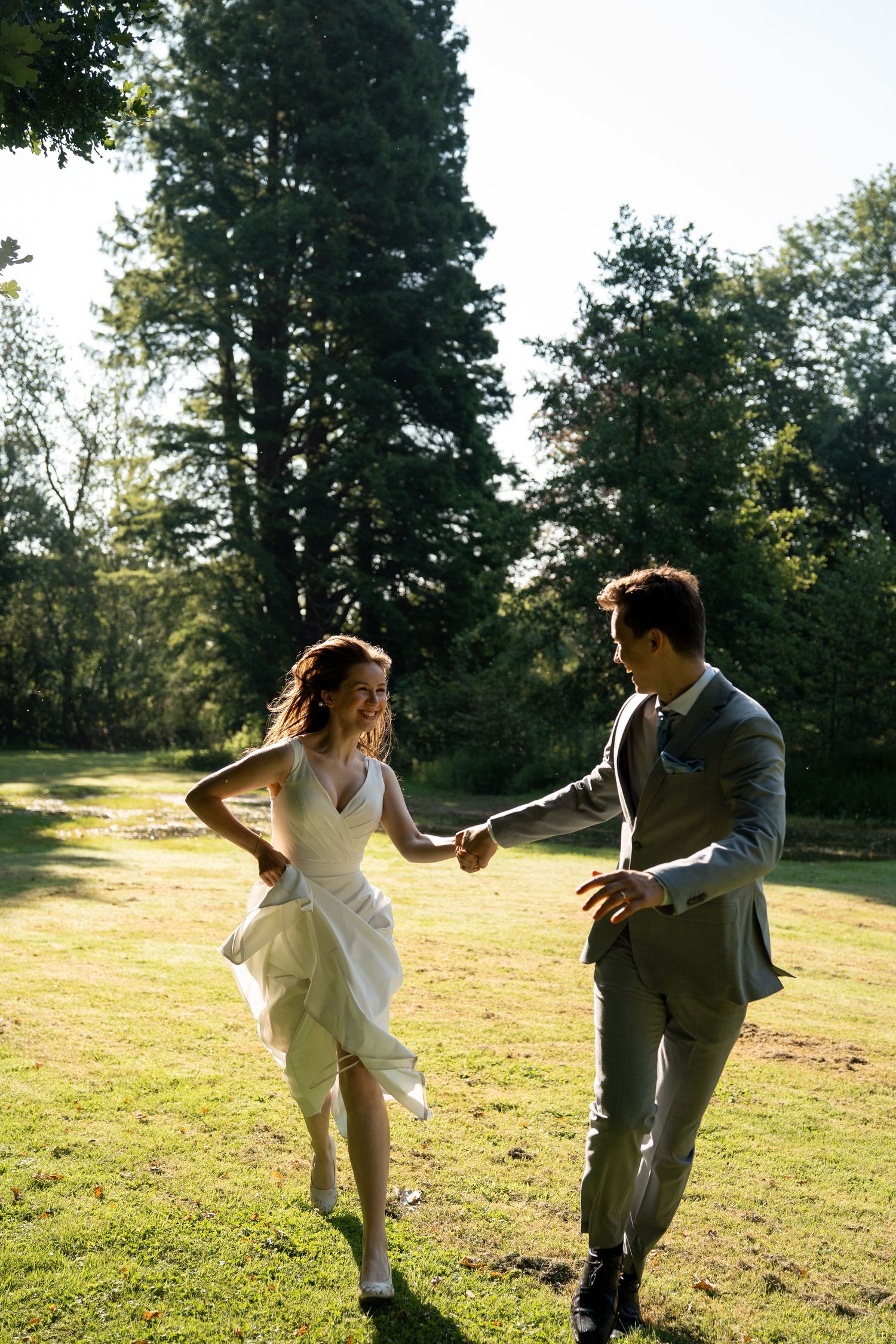 Joyful bride and groom holding hands and dancing on a sunny green lawn surrounded by tall trees.