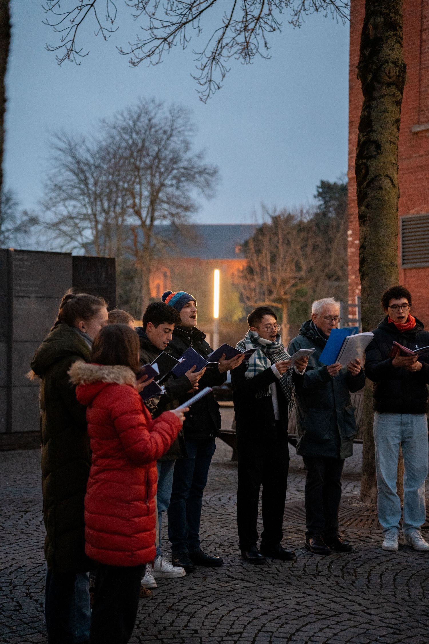 Group of carol singers holding songbooks and performing outdoors near a brick church at dusk.