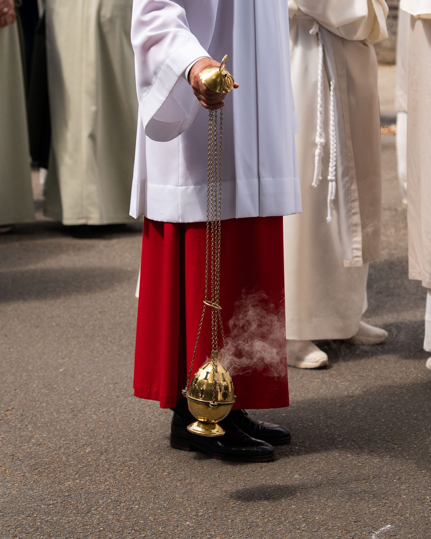 Altar server in white and red robes swinging a golden thurible with incense smoke during a religious procession.