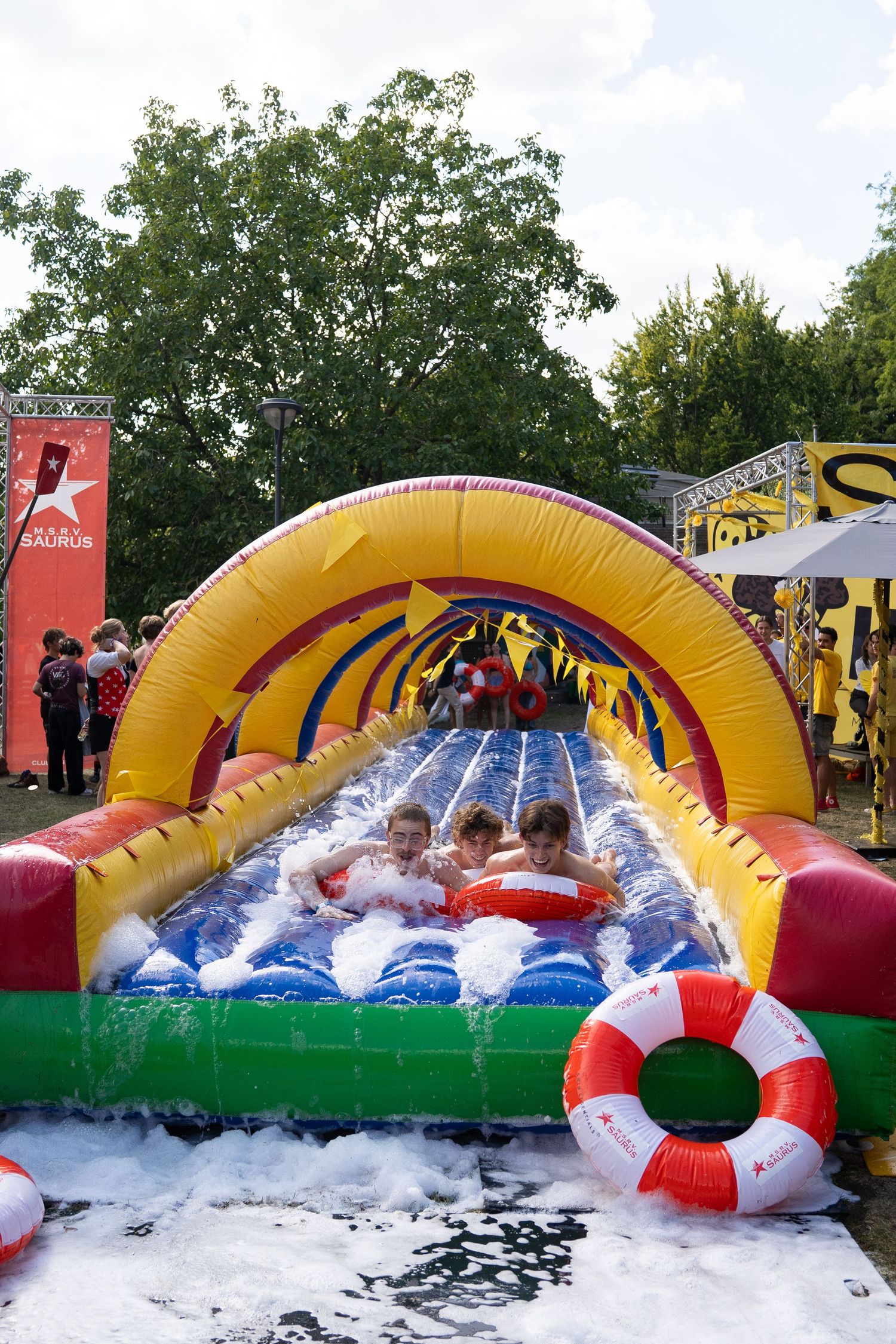 Children sliding down a foam-filled inflatable water slide at a vibrant outdoor summer festival.