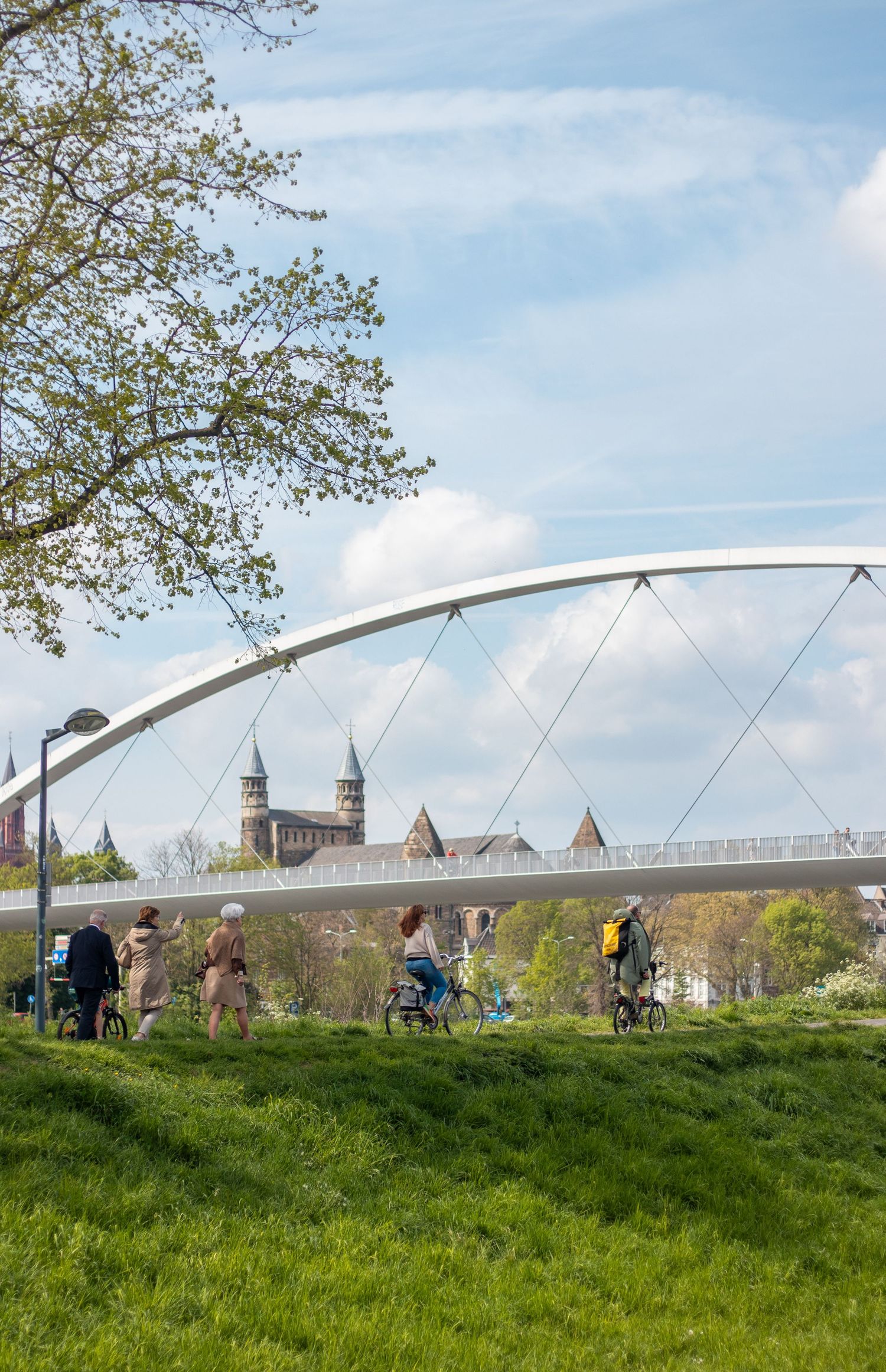 People cycling and walking across a modern white arch bridge over the Maas river in Maastricht in spring.