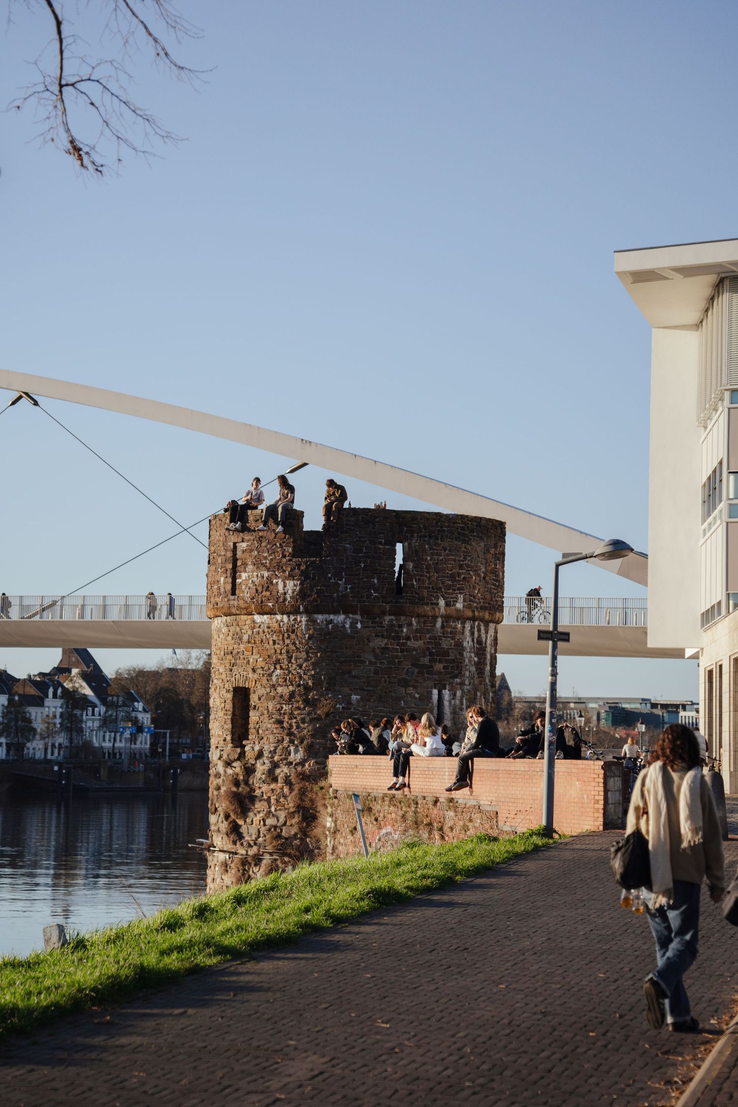 People atop a historic stone tower by a river with a modern pedestrian bridge in the background.