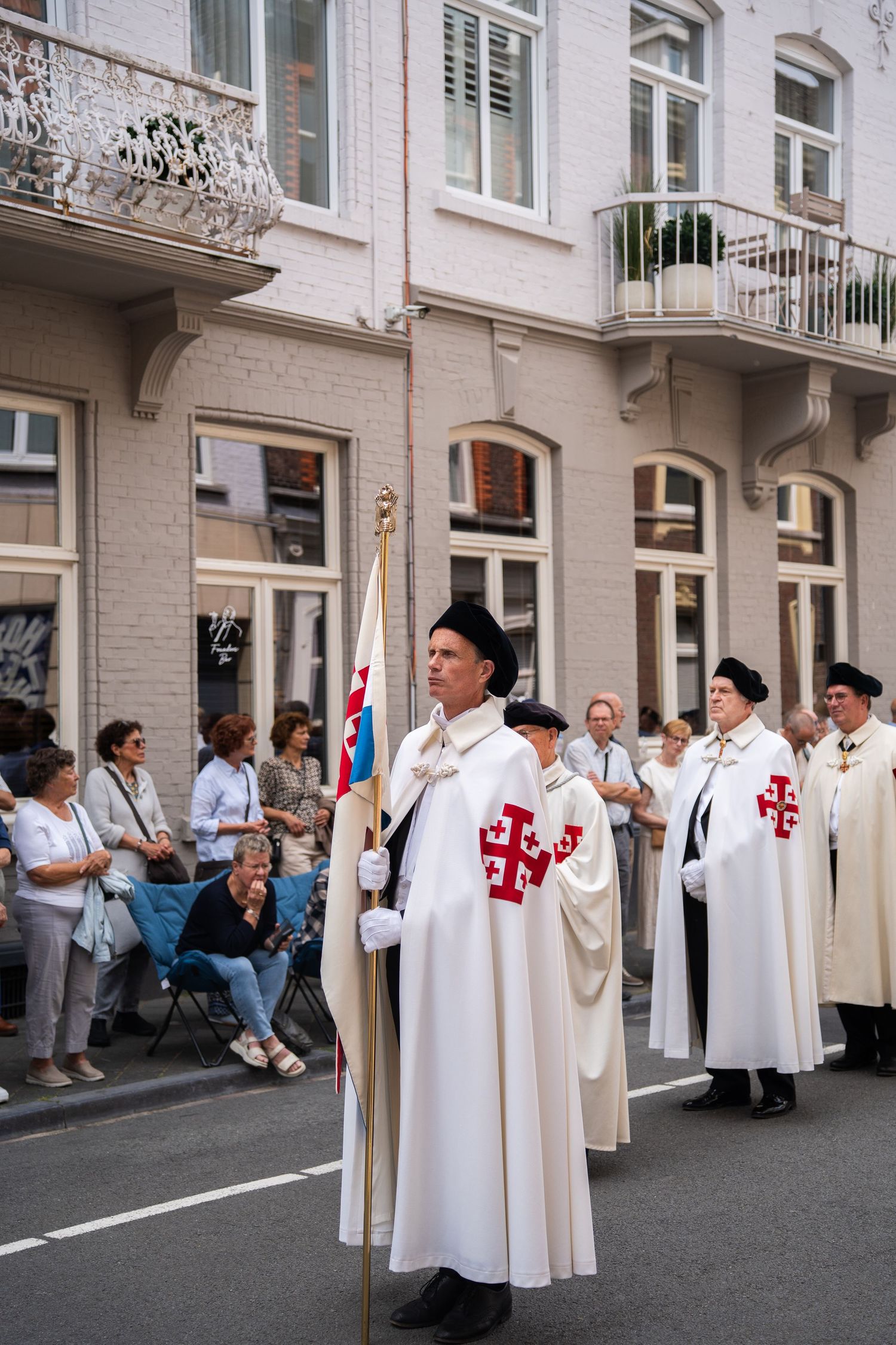 Knights in white robes with red Jerusalem crosses carry flag in a solemn religious procession.