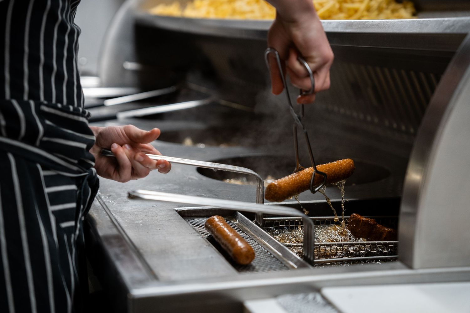 Chef frying sausages in commercial deep fryer using tongs in professional kitchen with striped apron.