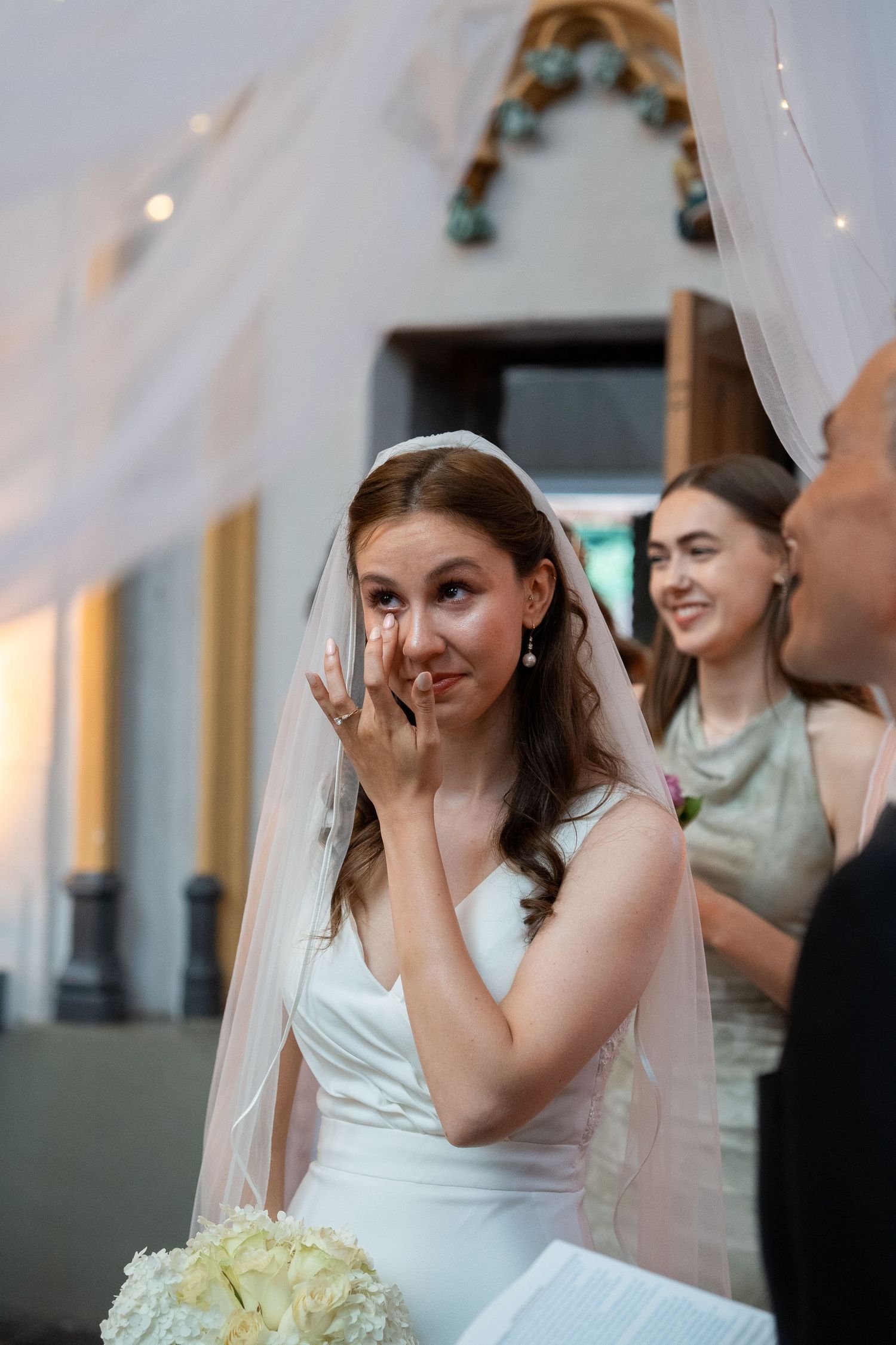 Emotional bride wiping tears during wedding ceremony with smiling bridesmaid behind her.