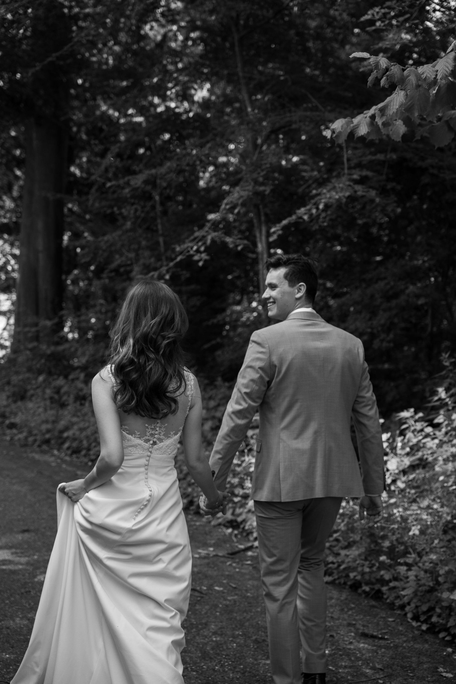 Black and white photo of bride and groom walking hand in hand along a lush forest path.