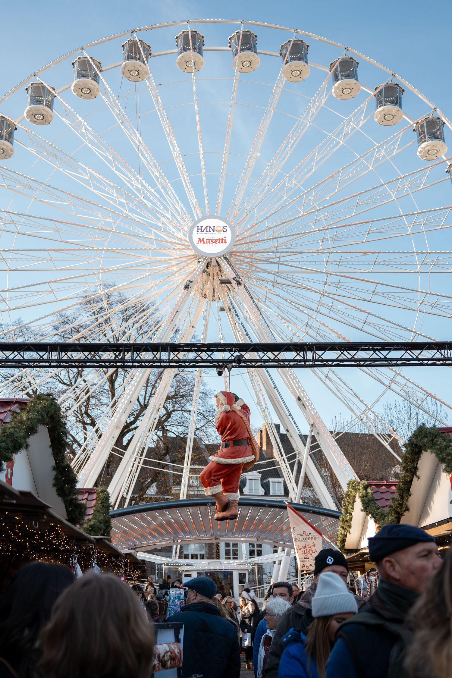 Festive Christmas market with a large white Ferris wheel and Santa Claus decoration under blue sky.