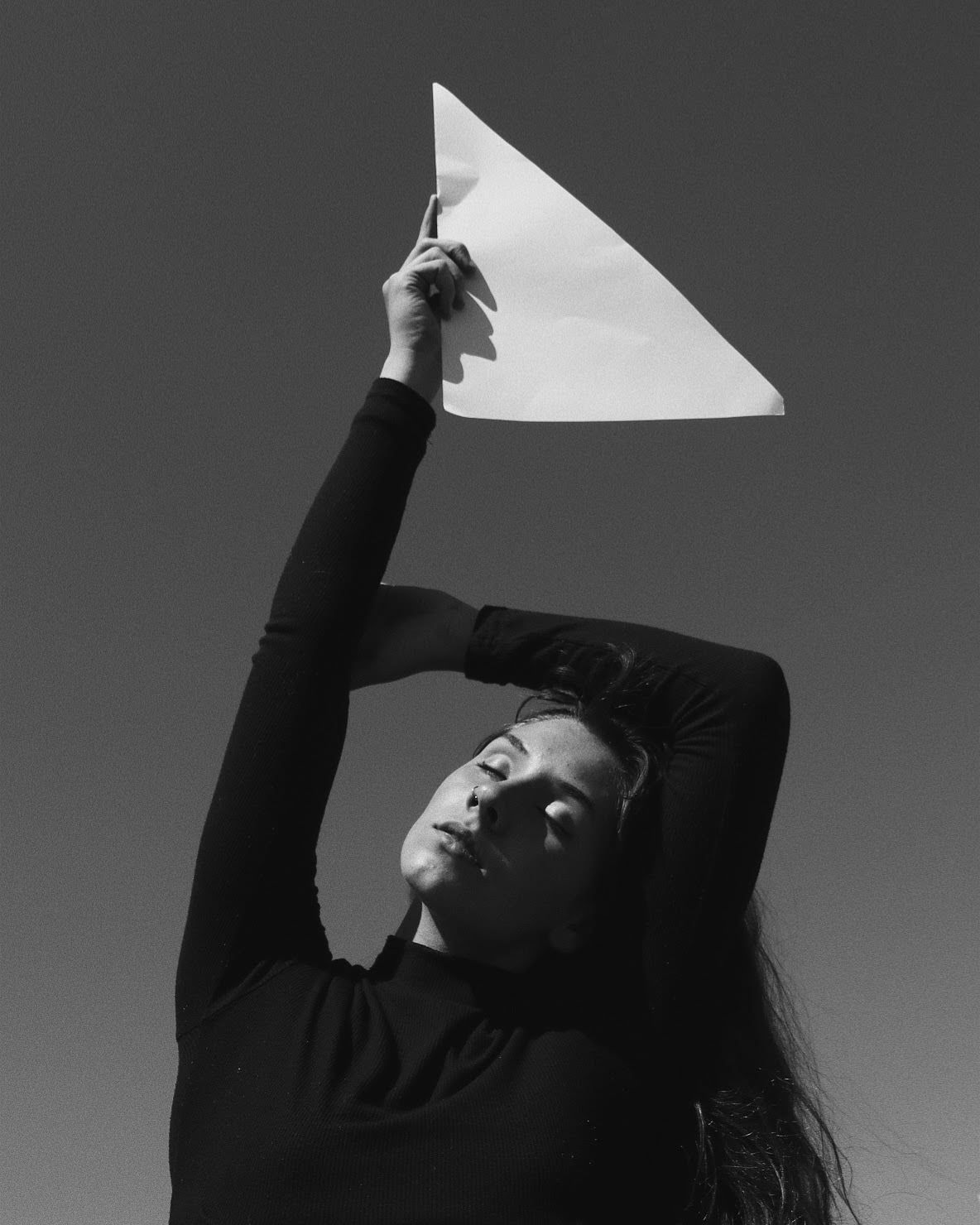 Black and white photo of woman holding paper airplane above head against clear sky.