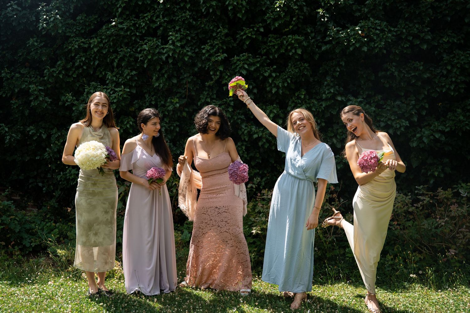 Five bridesmaids in mismatched pastel dresses holding colorful bouquets while posing outdoors in sunlight.