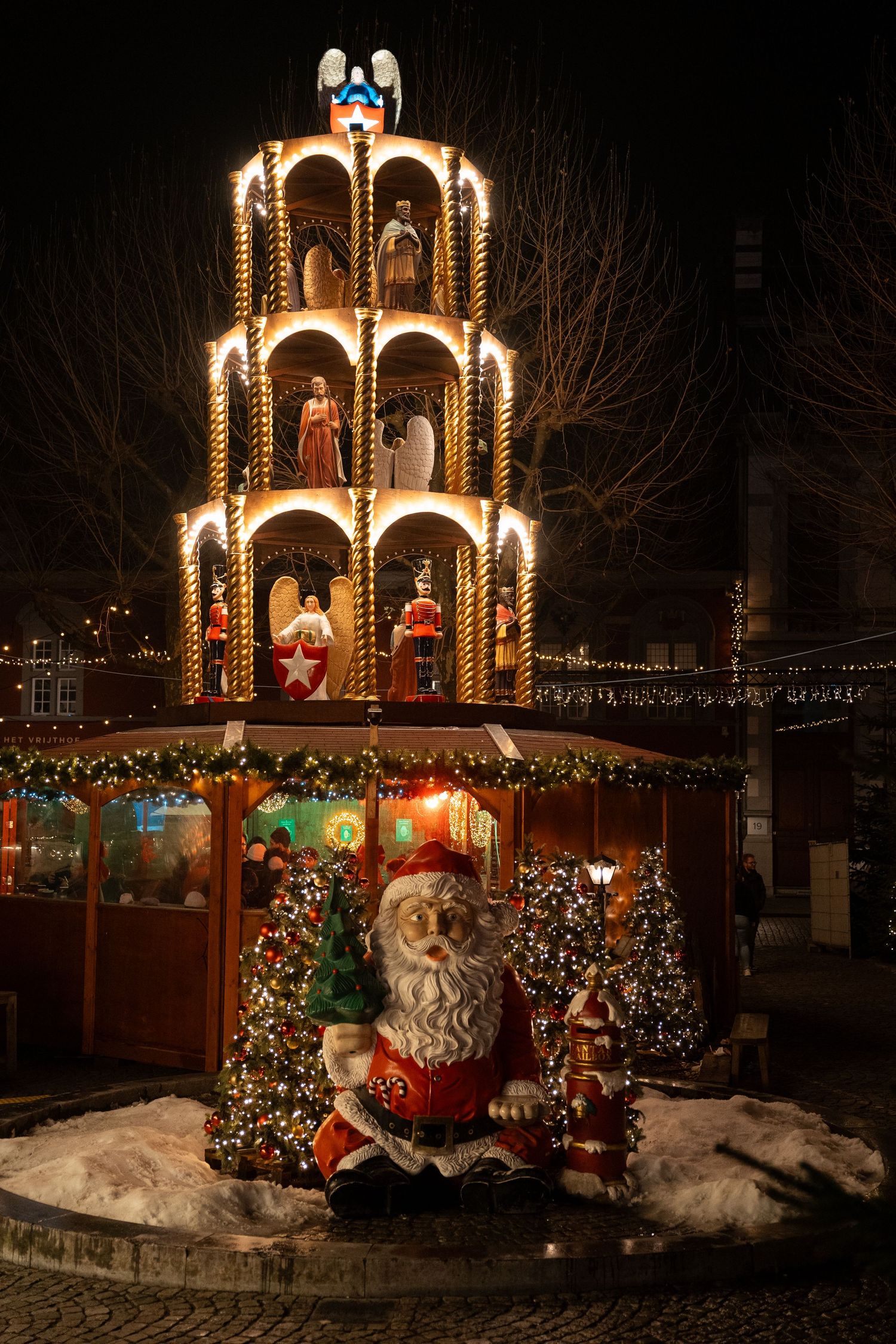 Illuminated multi-tiered Christmas pyramid with nativity figures and a Santa Claus statue at a winter market.