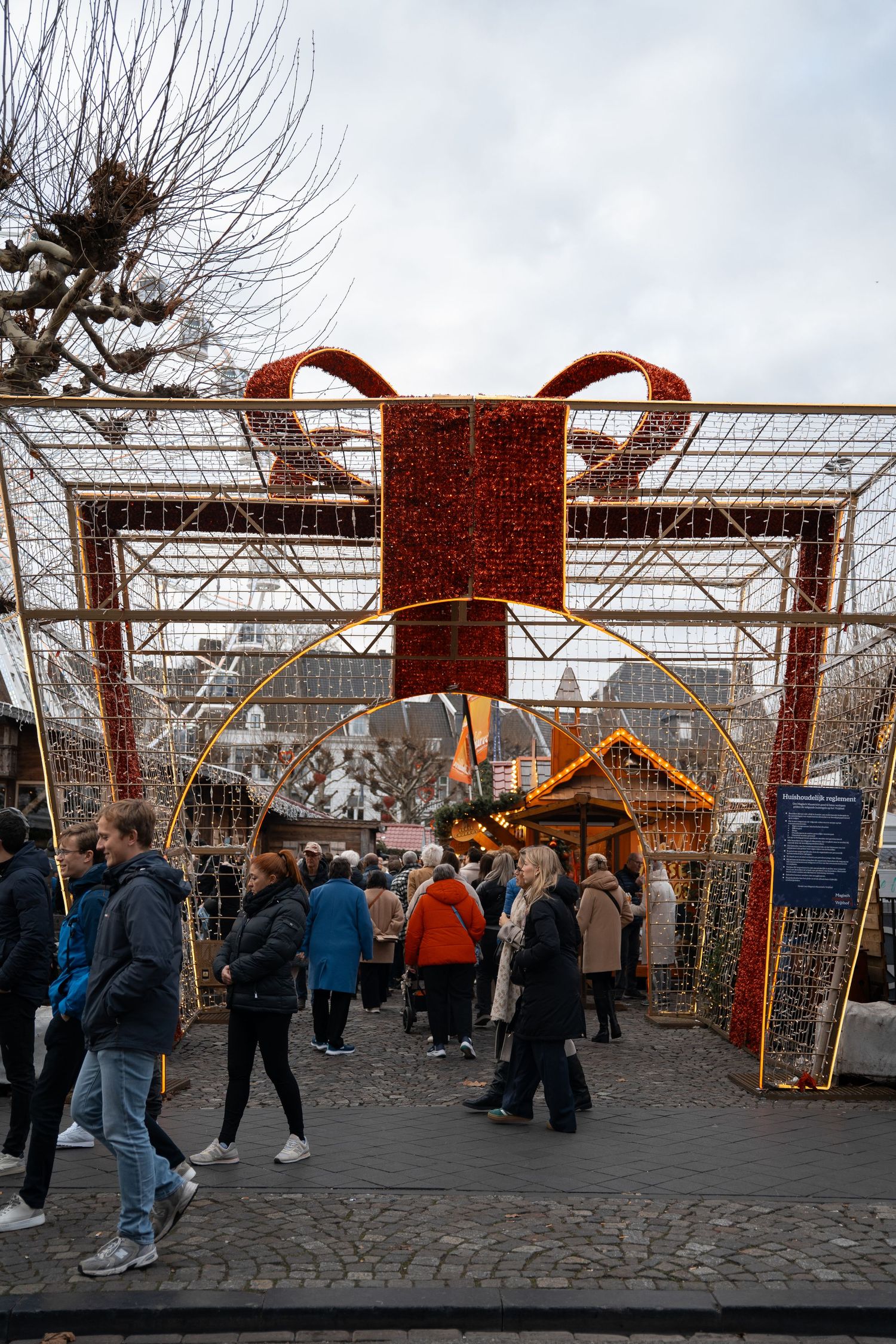 Visitors walking through a large golden Christmas gift box gate decoration at a festive market.