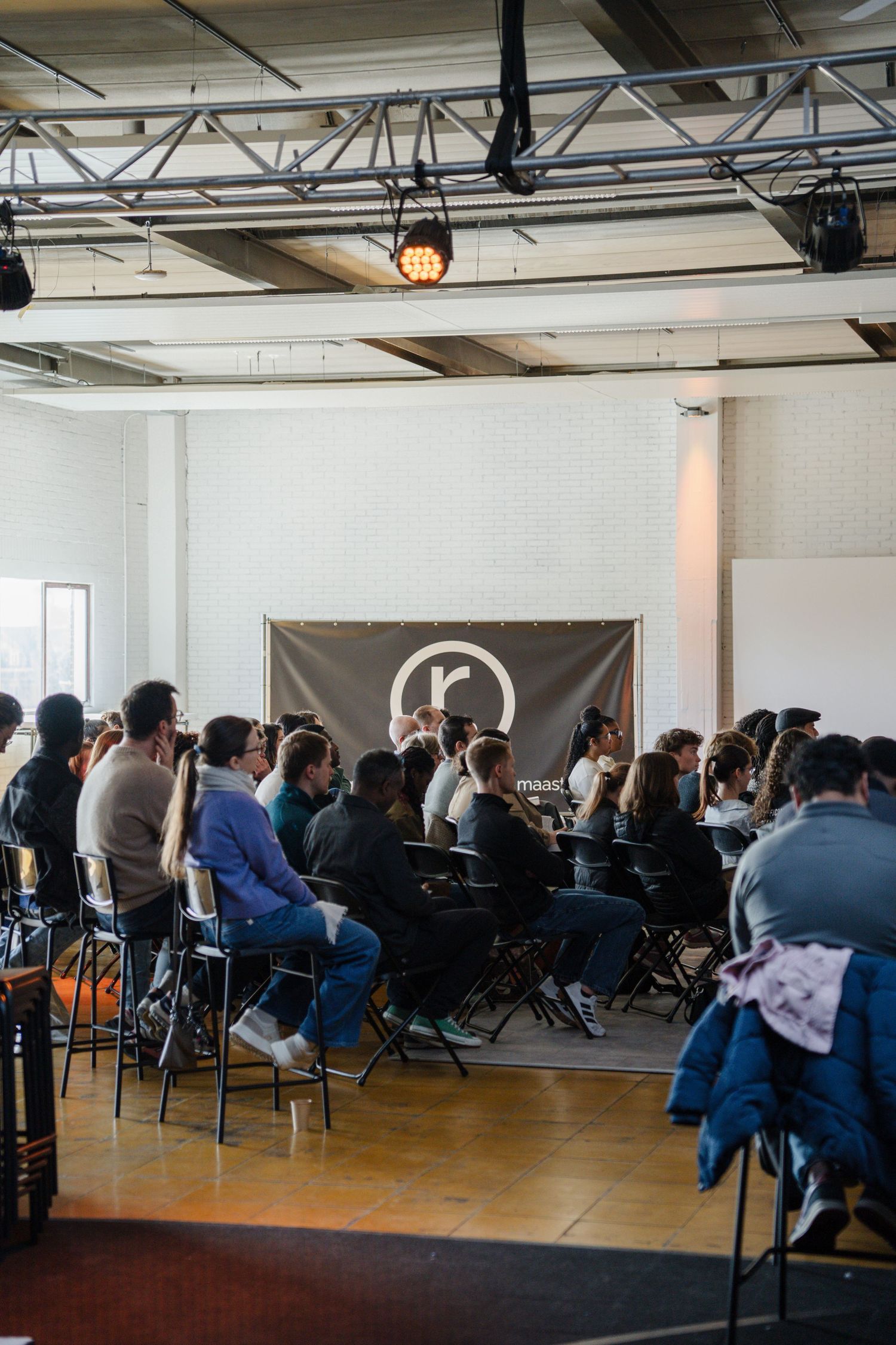 Audience seated in chairs at an indoor church gathering with a speaker and a banner logo on the wall.