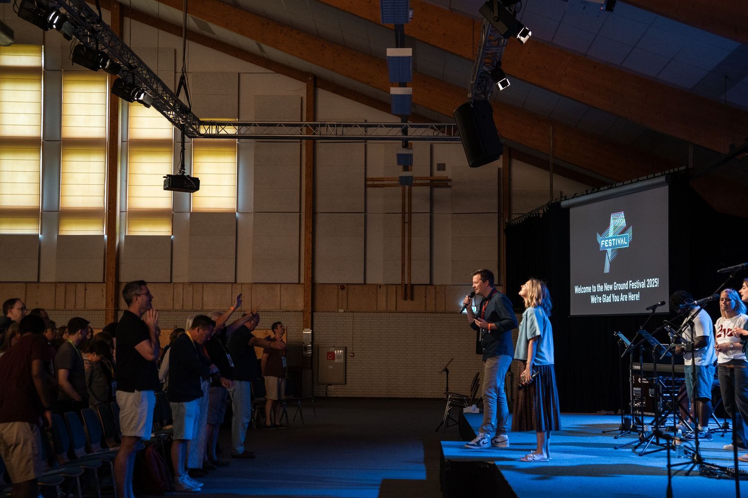 Worship leader on stage with congregation raising hands during a church service with projection screen.