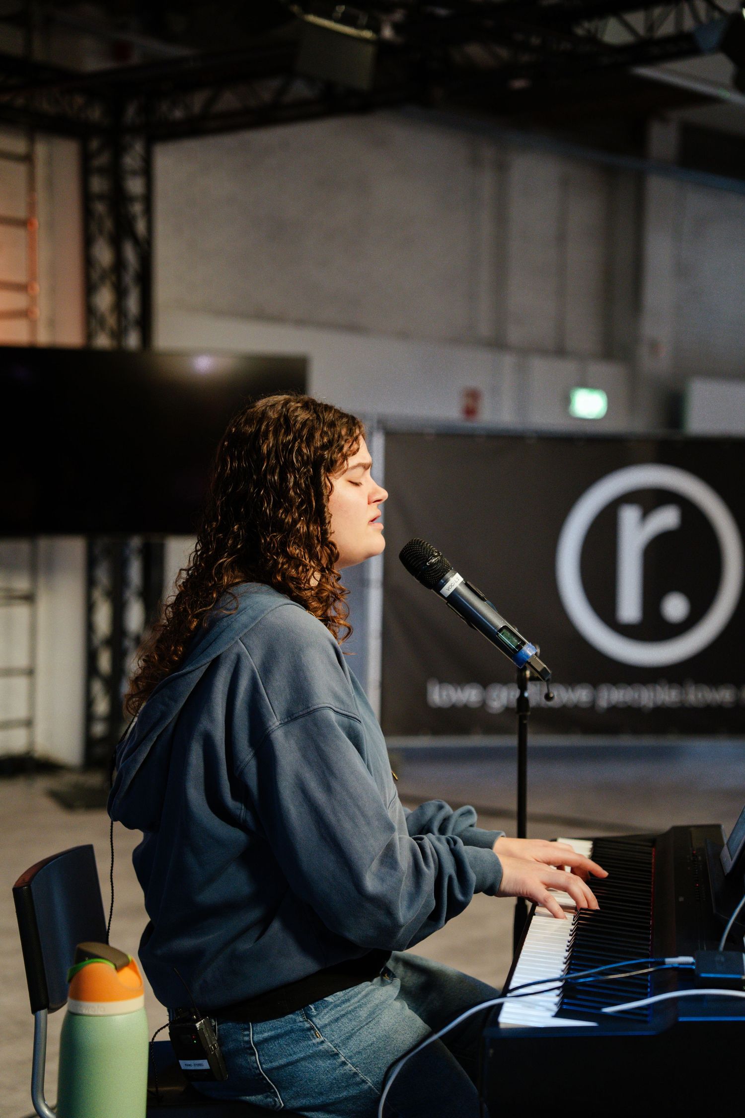 Curly-haired woman plays piano and sings at microphone during worship service with logo banner.