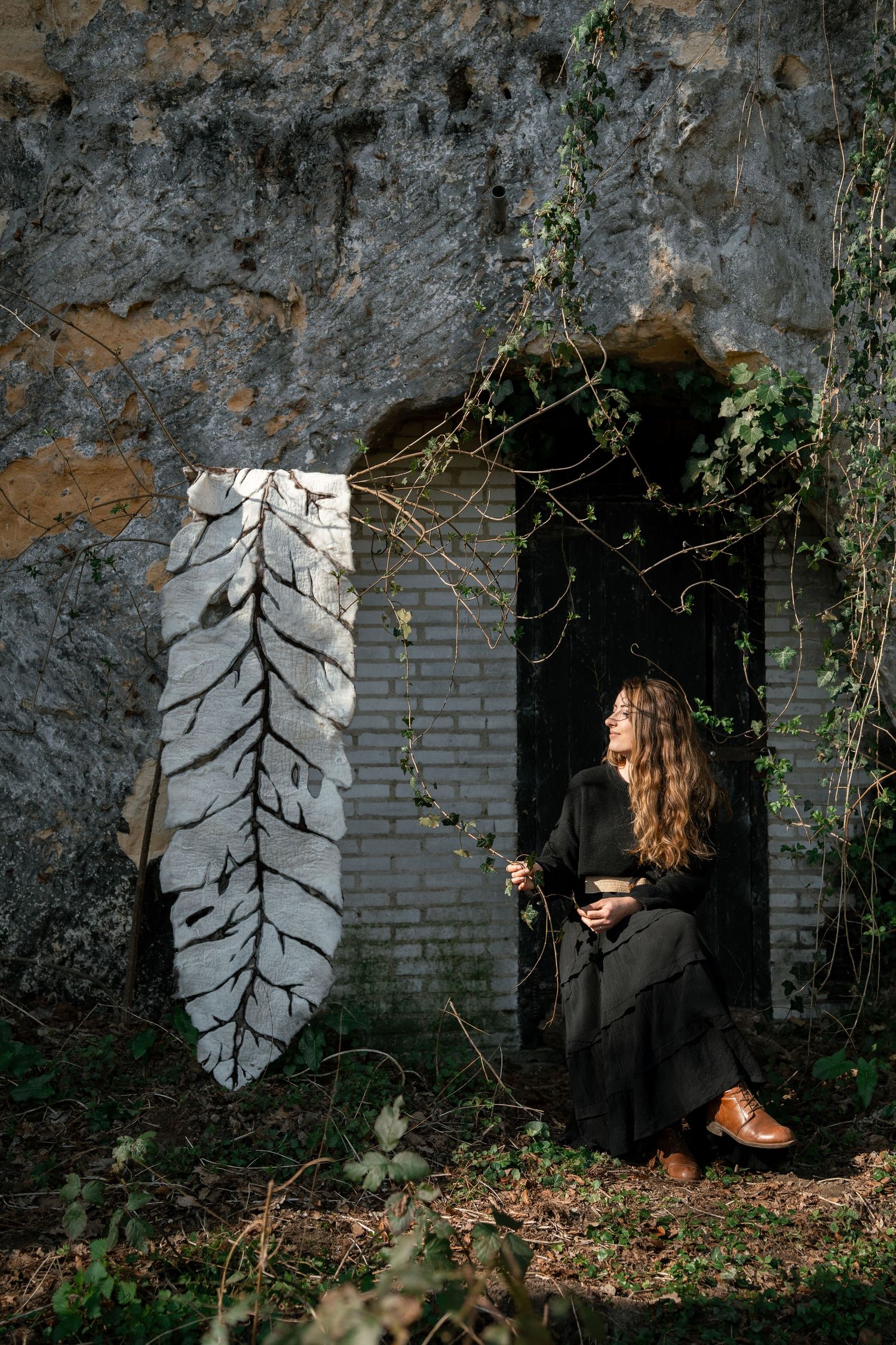 Woman in black dress seated in a stone doorway beside a large white felted leaf artwork.
