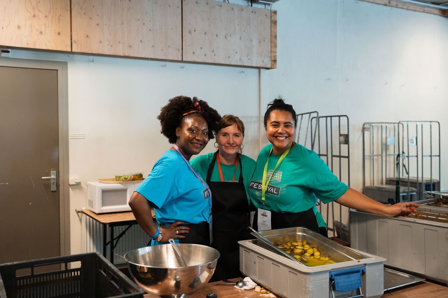Three smiling women posing together in a kitchen while preparing food at a catering event.