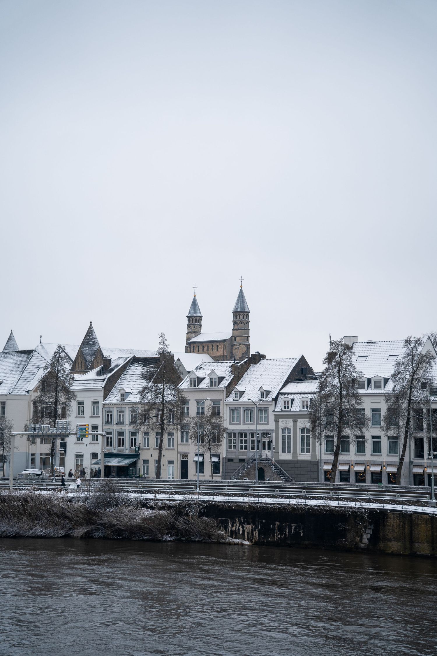 Snow-covered historic European buildings along a river waterfront with twin church towers in winter.