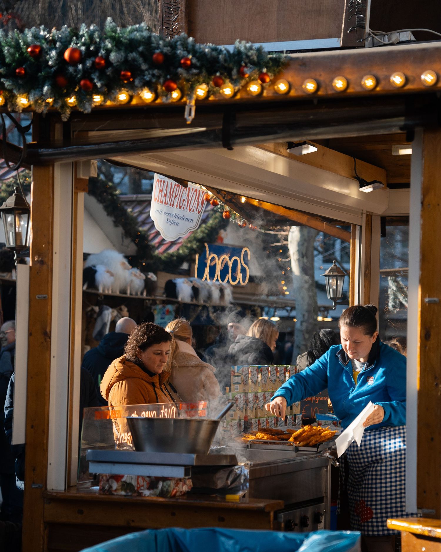 Two vendors cooking and serving food at a warmly lit Christmas market stall with festive decorations.