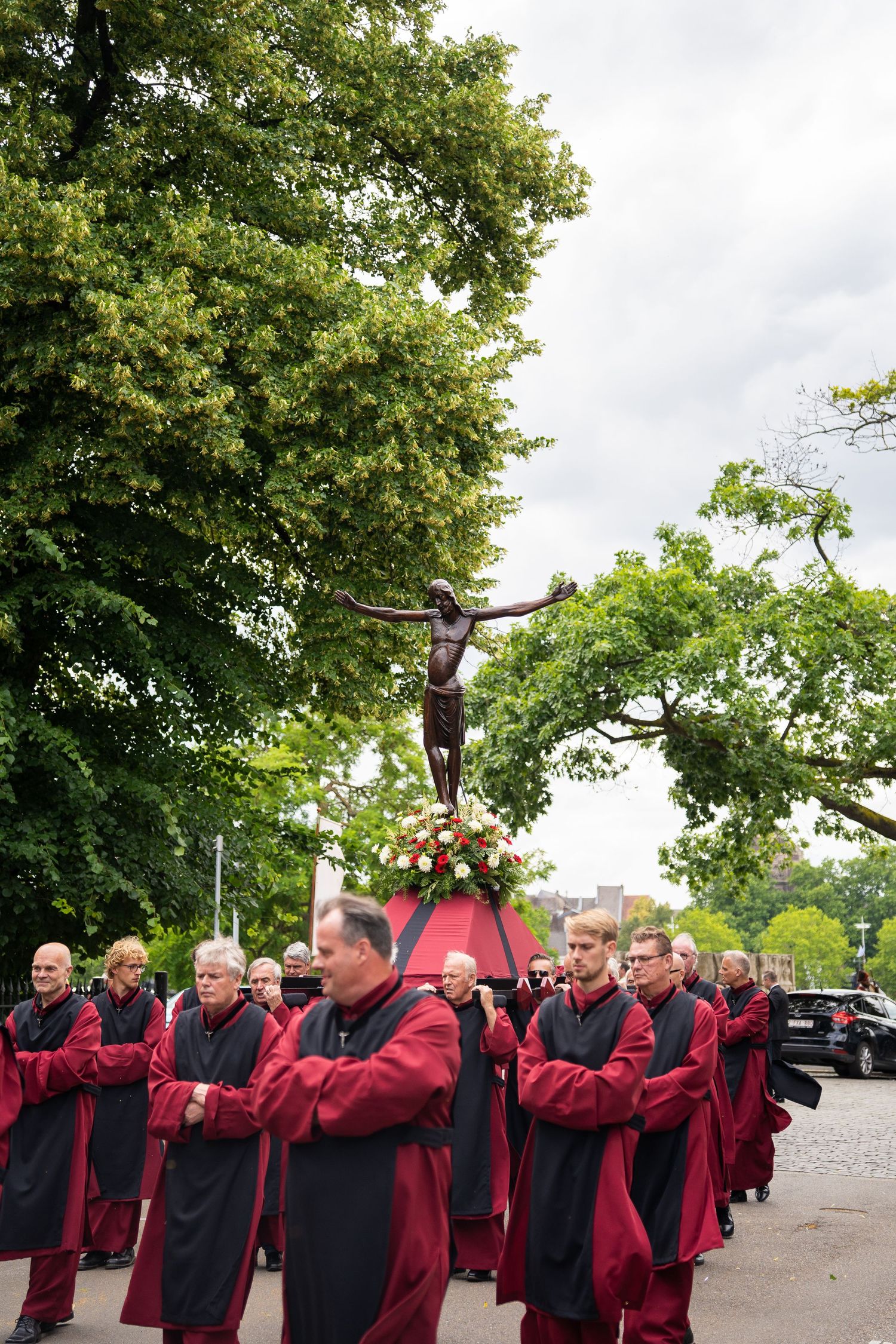 Procession of clergy in dark red robes carrying a crucifix statue adorned with flowers through a street.