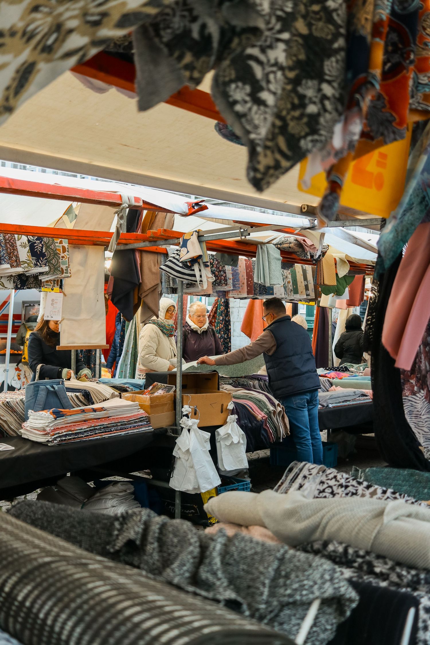 Busy outdoor textile market stall with vendors and customers browsing fabrics.