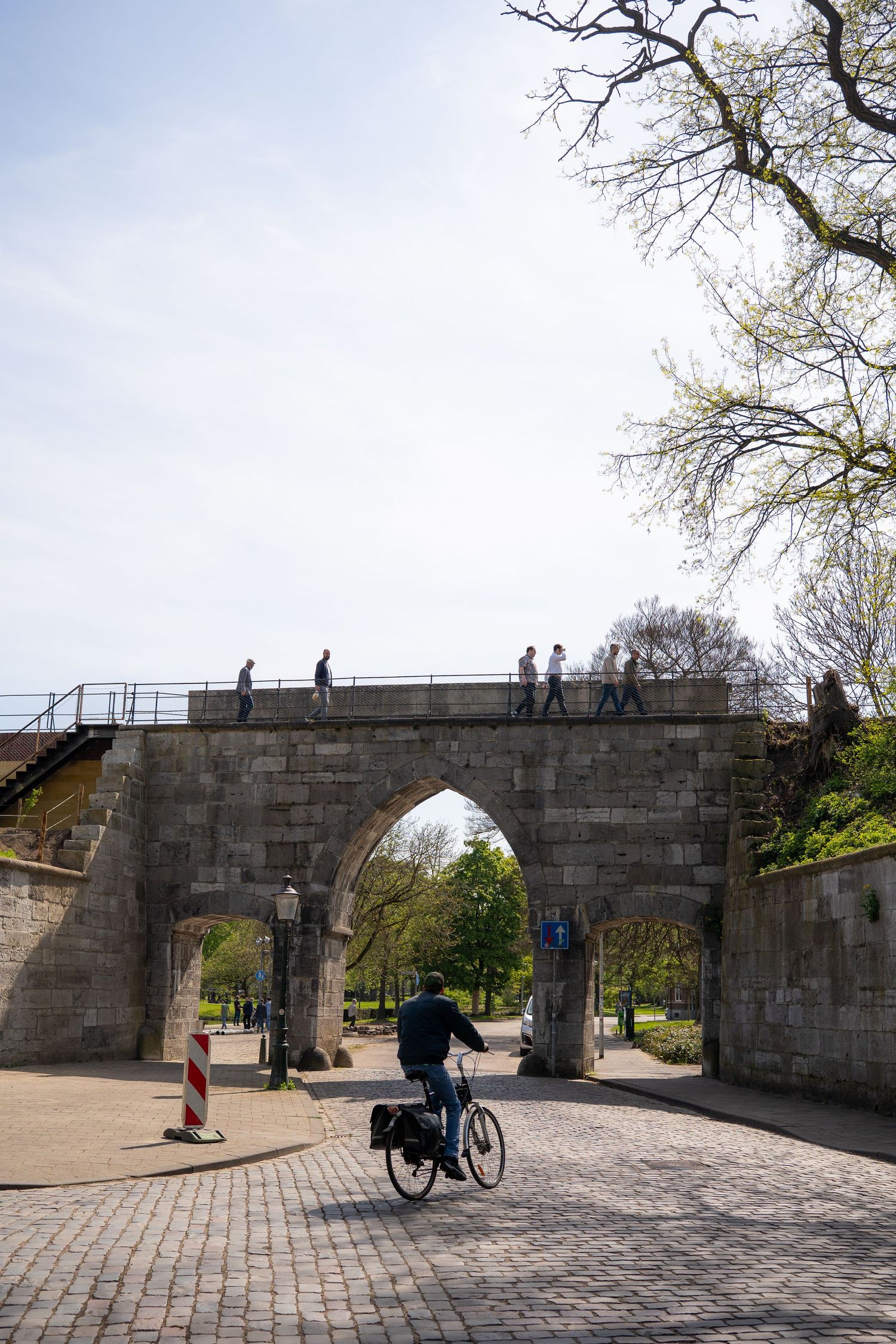 Cyclist passing through a historic stone arch bridge gateway in a European city park on a sunny day.