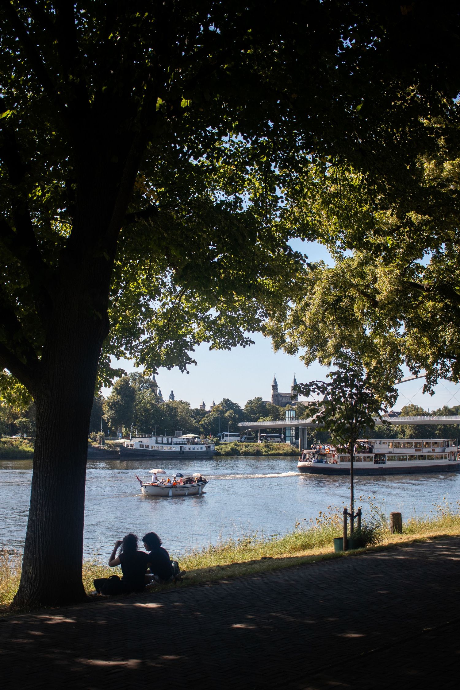 Two people sitting under a large tree by a river watching boats pass with a cathedral in the distance.