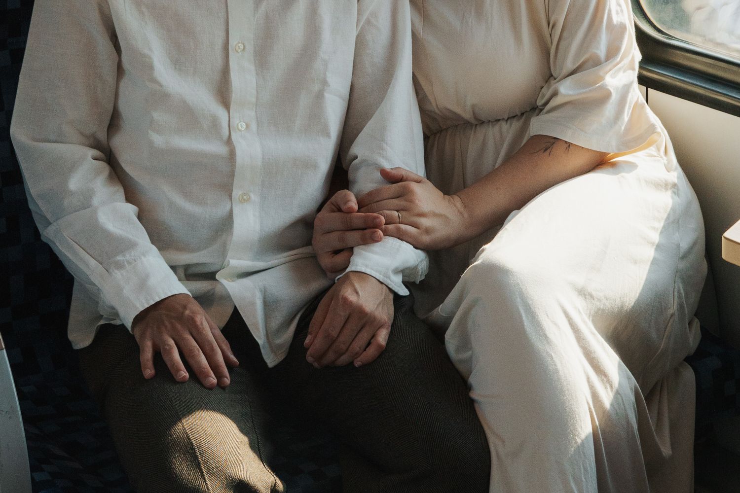 Couple in white attire sitting together holding hands, wedding rings visible, romantic and intimate moment.
