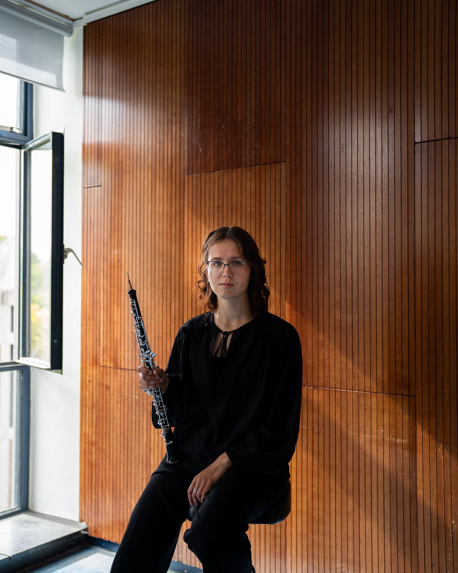 Female oboist in black top seated beside a wooden wall holding her oboe near a bright window.