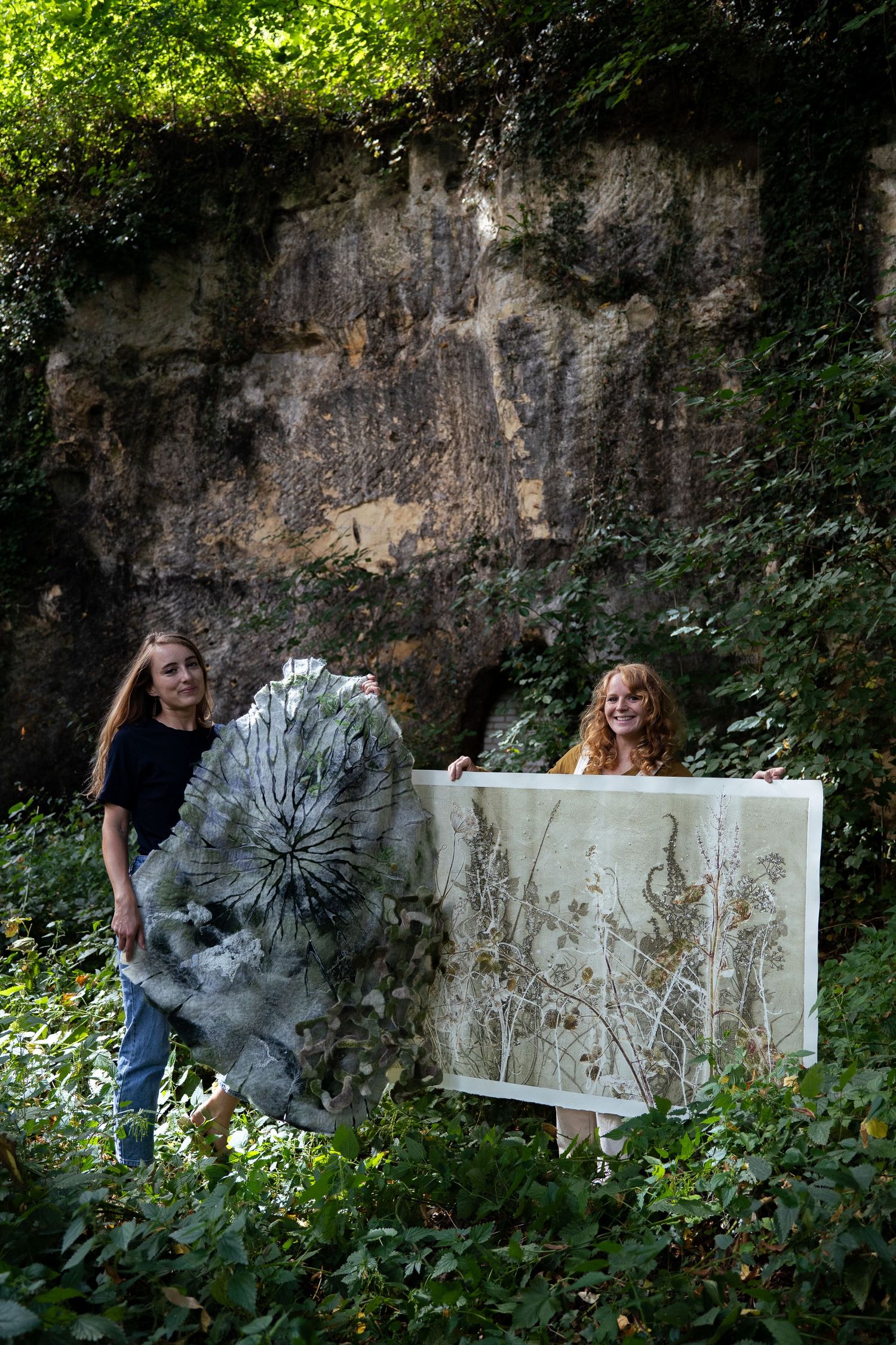 Two women holding botanical art pieces with pressed plants in lush green surroundings near rock face.