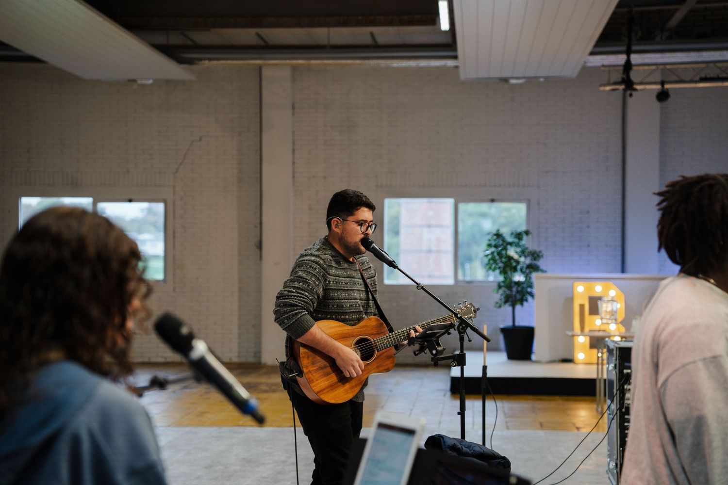Male guitarist with glasses performs at microphone during casual church worship rehearsal session.