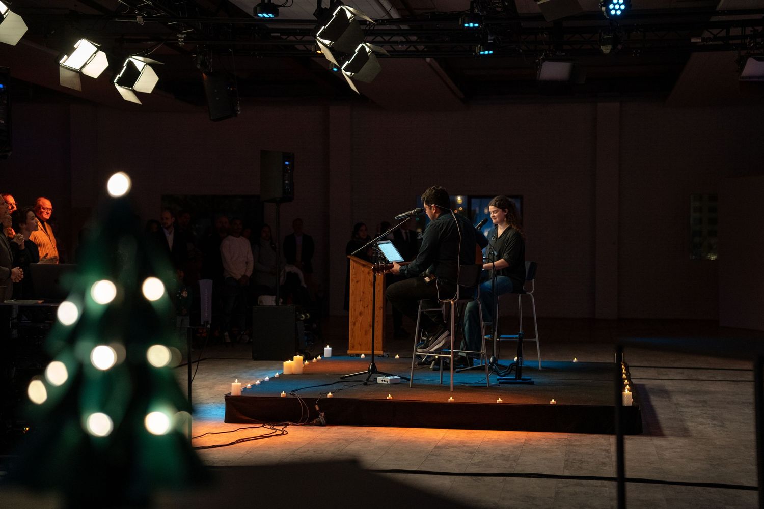 Two speakers on a dimly lit stage at a podium during an intimate church service with Christmas decor.
