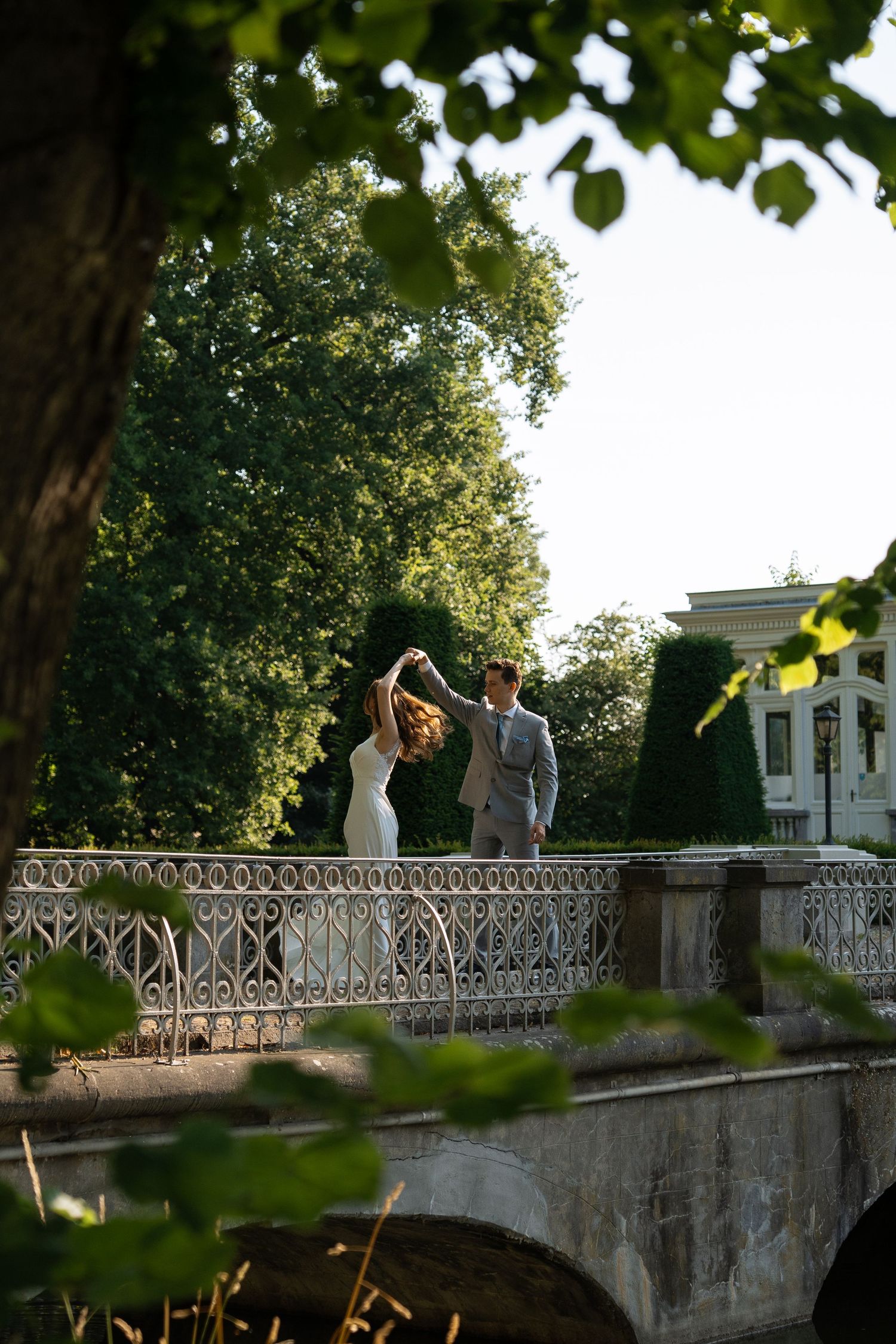 Bride and groom dancing on an ornate bridge in a lush green park setting.