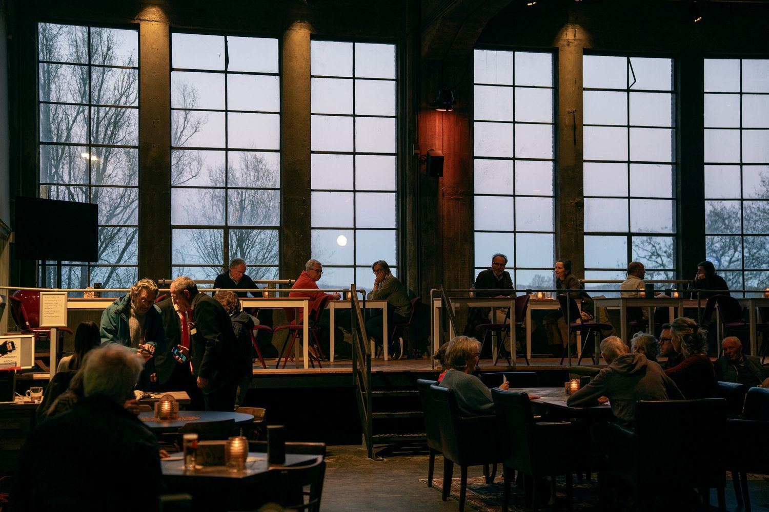 People sit at candlelit tables in an industrial venue with large warehouse windows at dusk.