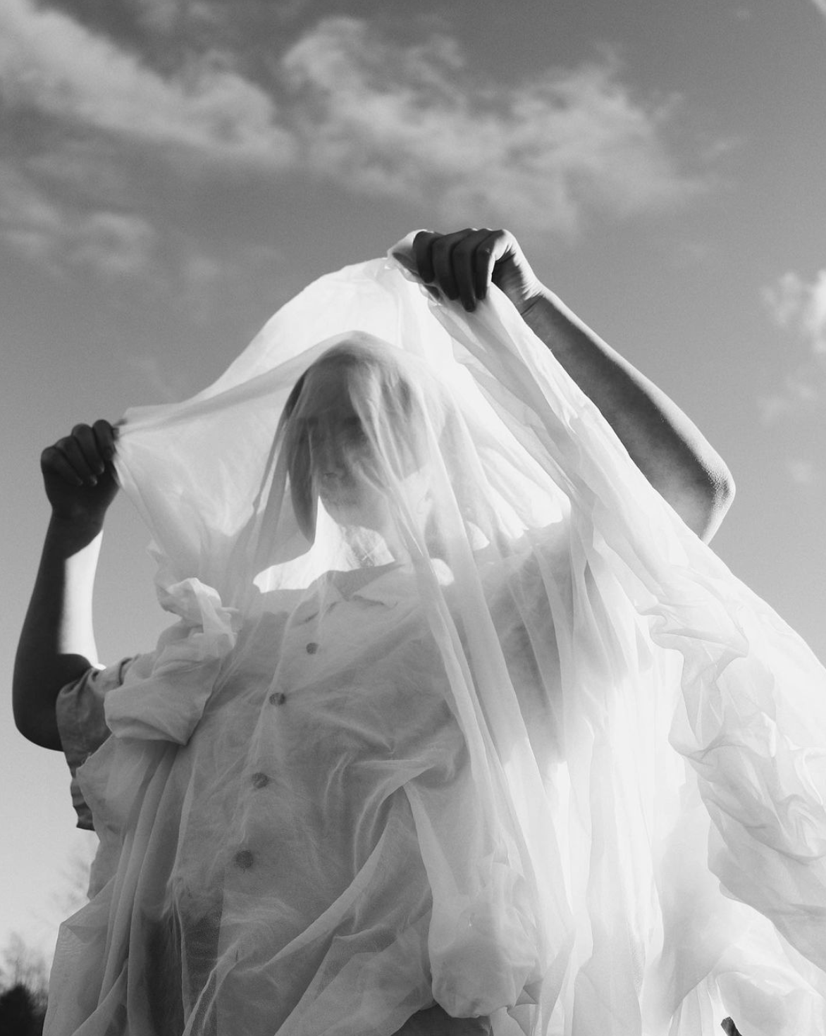 Black and white photo of two people lifting a sheer veil fabric against a cloudy sky outdoors.